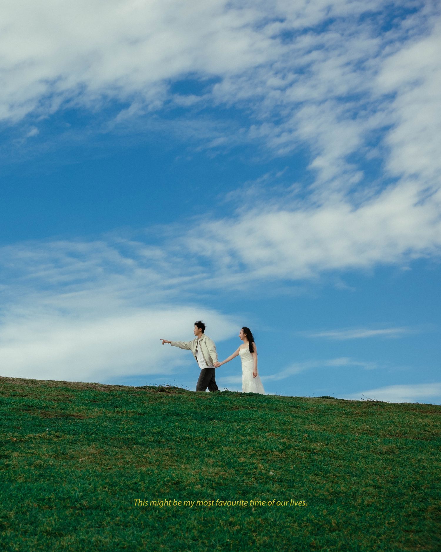 Couple holding hands on a lush green hill under a bright blue sky with scattered white clouds.