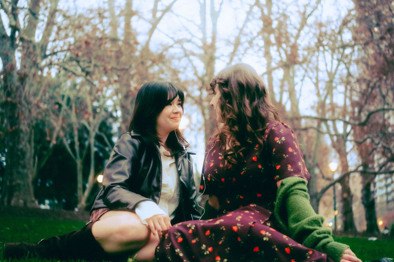 Two young women sitting on grass in a park, smiling at each other among bare autumn trees.