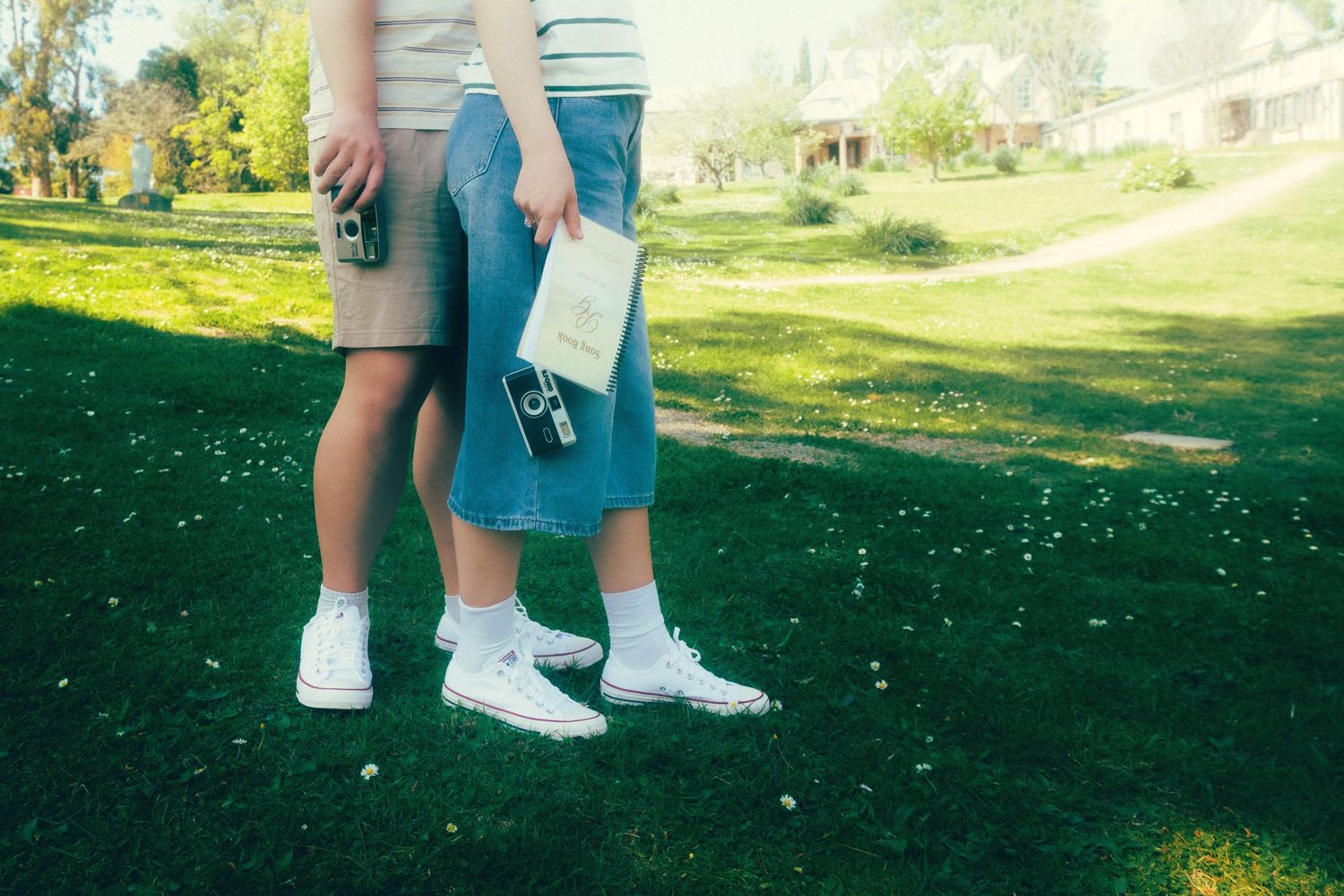 Two people standing on green grass wearing white sneakers, shorts, and skirts, holding a camera and papers outdoors.