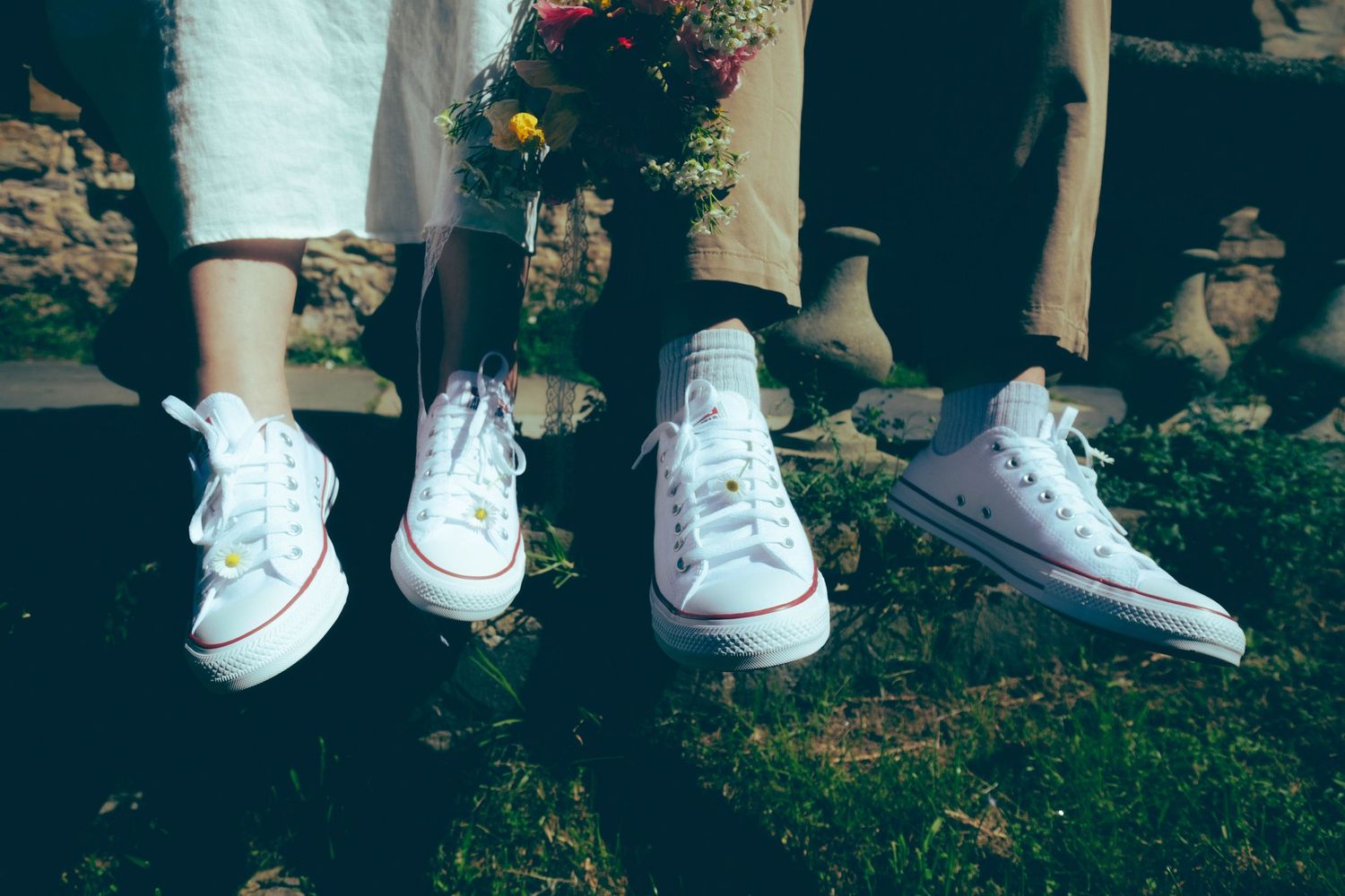 Bride and groom wearing matching white Converse sneakers on their wedding day, standing on green grass.