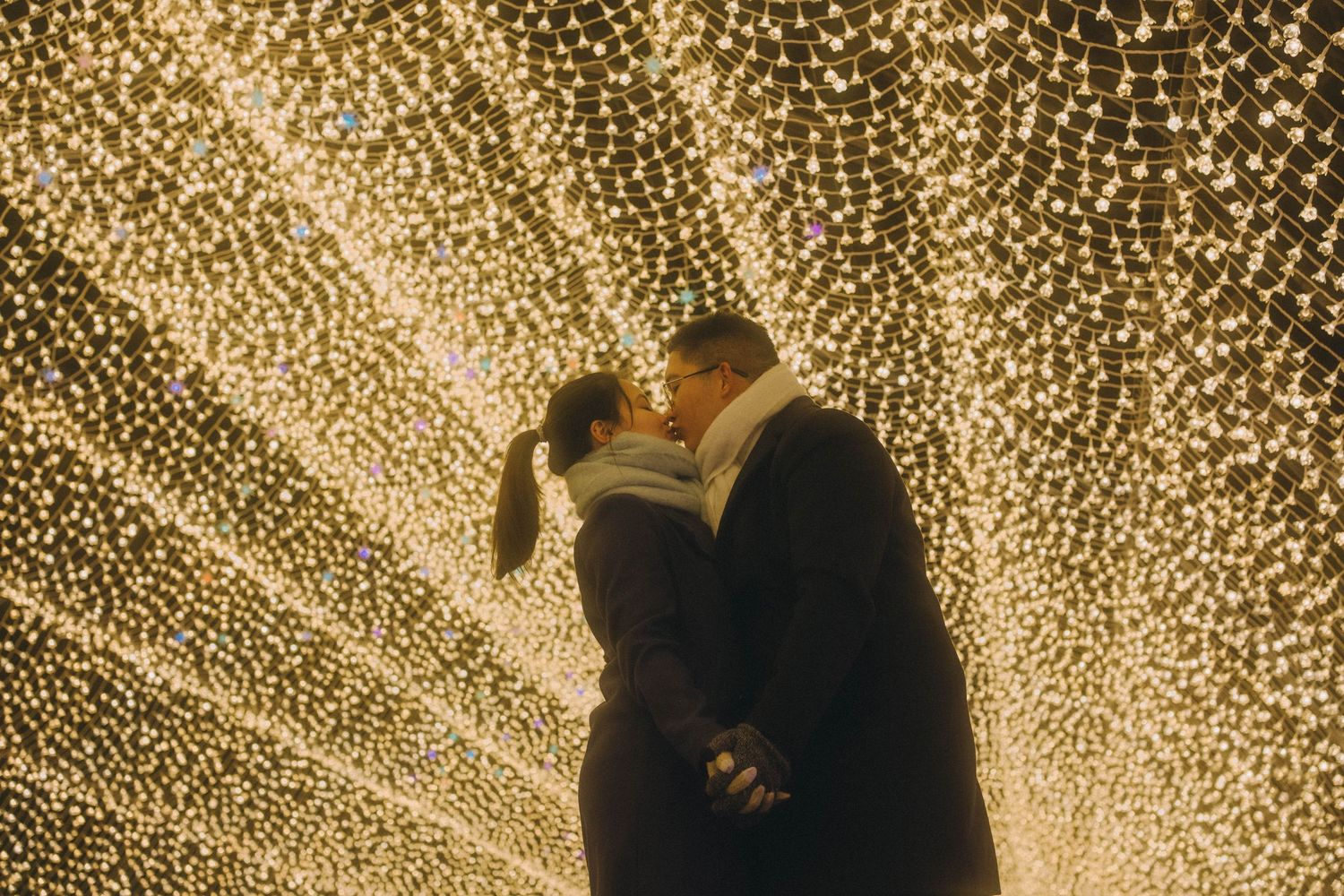 Romantic couple kissing under a canopy of warm glowing fairy lights at night.
