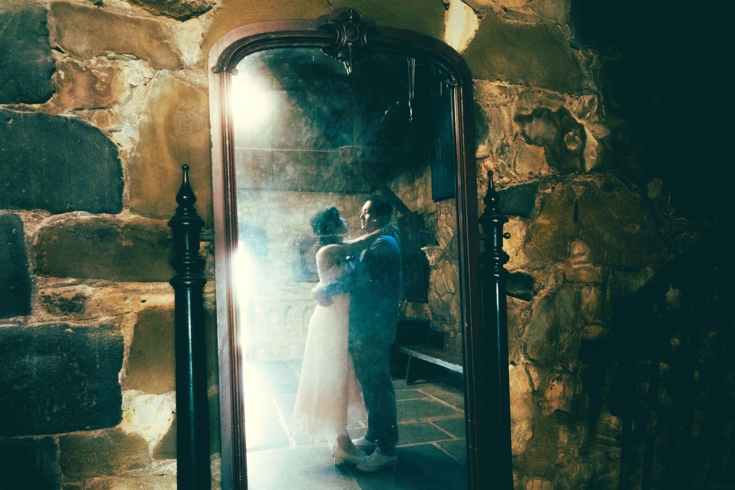 Couple sharing romantic kiss reflected in ornate mirror against rustic stone wall with dramatic lighting.