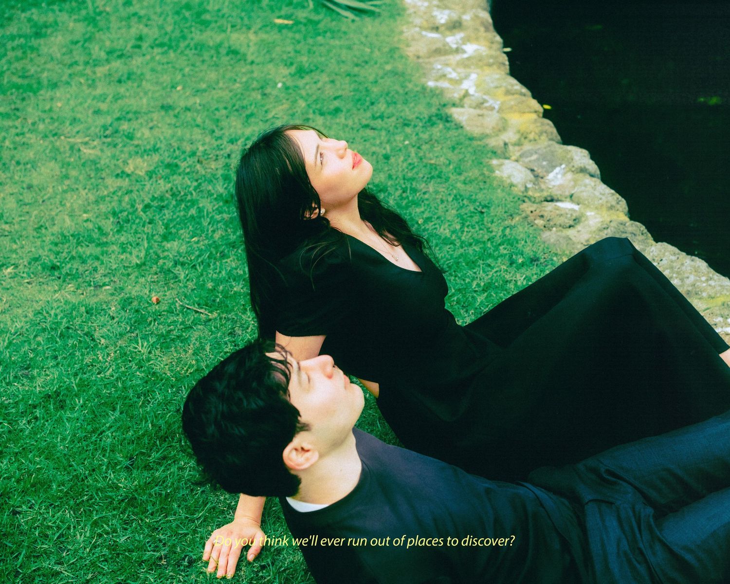 Two people in black clothing lying on green grass near water, gazing upward with text overlay about discovery.