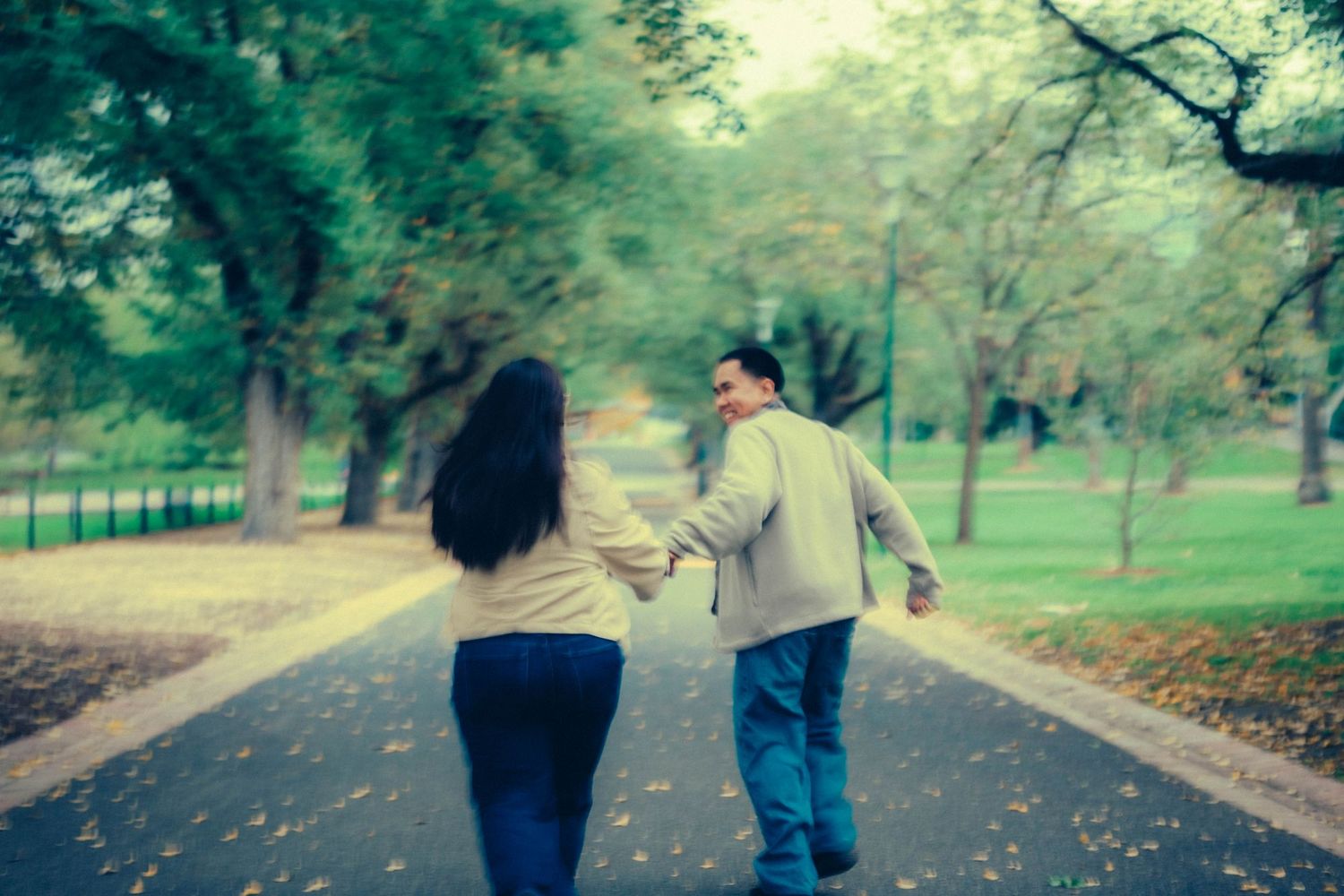 Couple holding hands walking down a tree-lined park path covered in fallen autumn leaves.