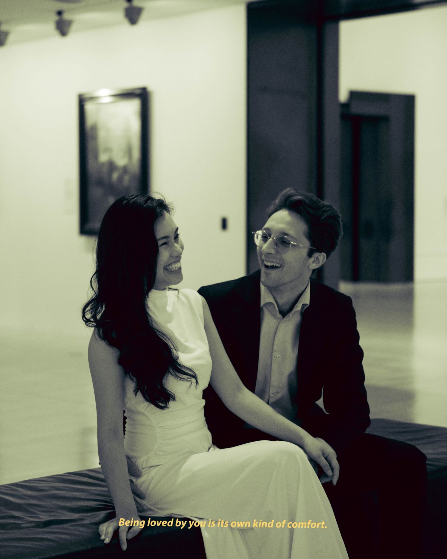 Elegant couple in black and white, sitting together in an art gallery, sharing a warm intimate smile.