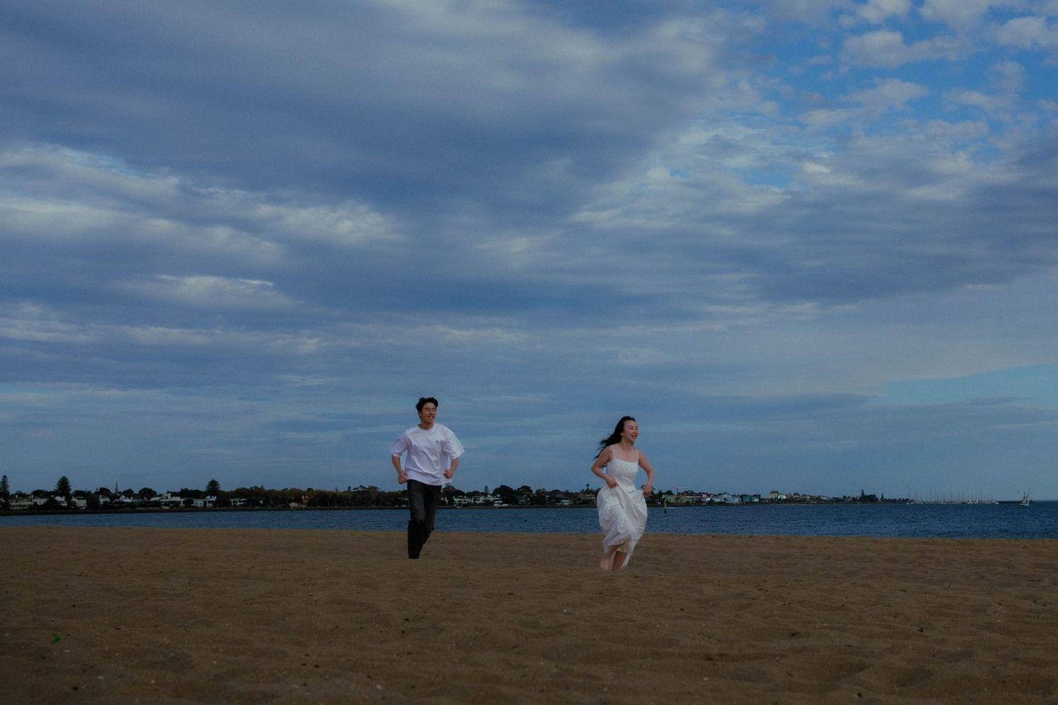 Couple running joyfully on a sandy beach under dramatic cloudy blue sky at dusk.
