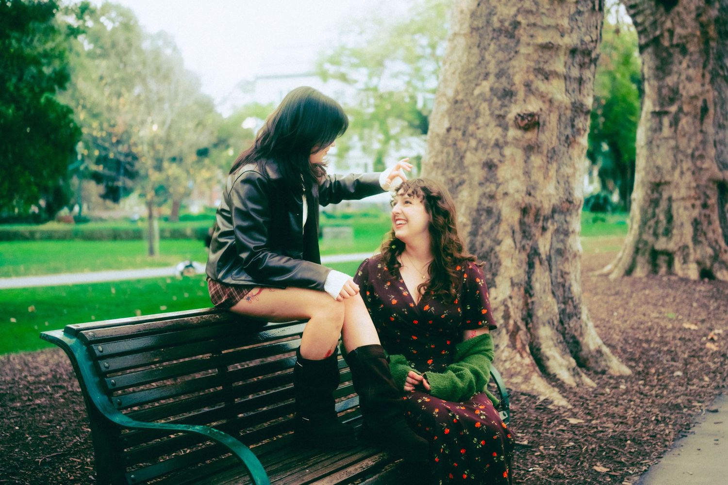Two women laughing together in a park, one sitting on a bench, sharing a joyful moment near a large tree.