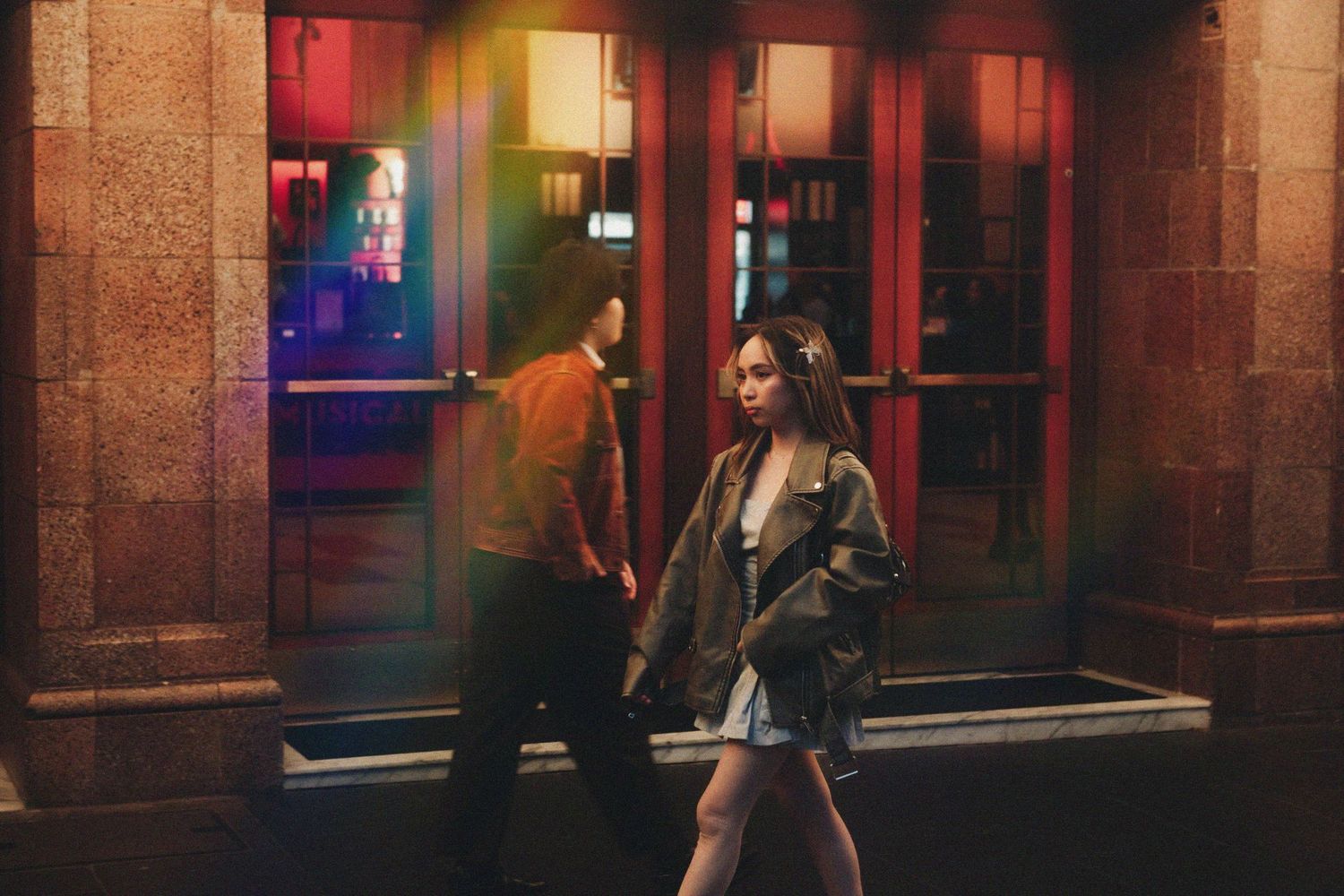 Young woman walking past a man outside a colorful illuminated building at night.