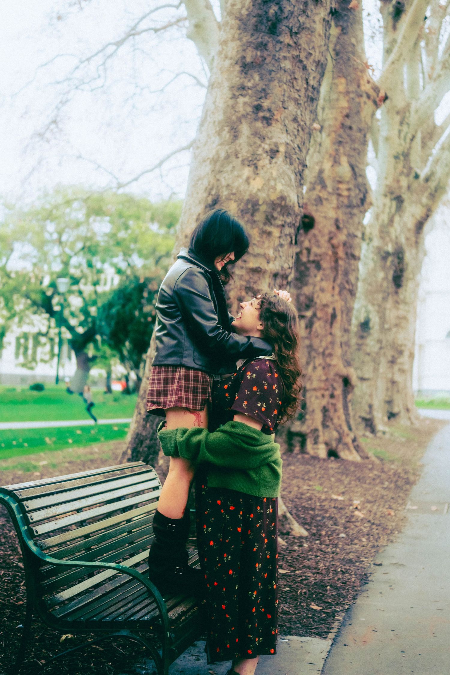 Two young women share a tender embrace near a park bench surrounded by large trees on an overcast day.
