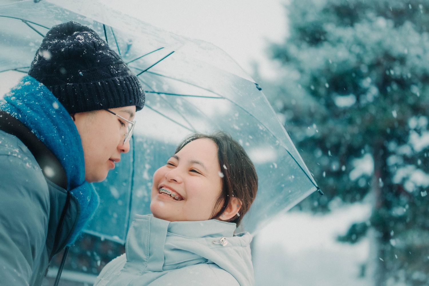 Smiling couple sharing a clear umbrella during gentle snowfall.