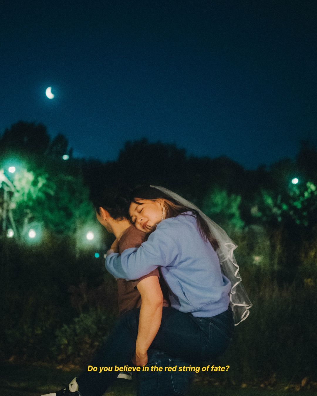 Woman resting her head on her fiance's shoulder under a moonlit night sky with glowing lights.