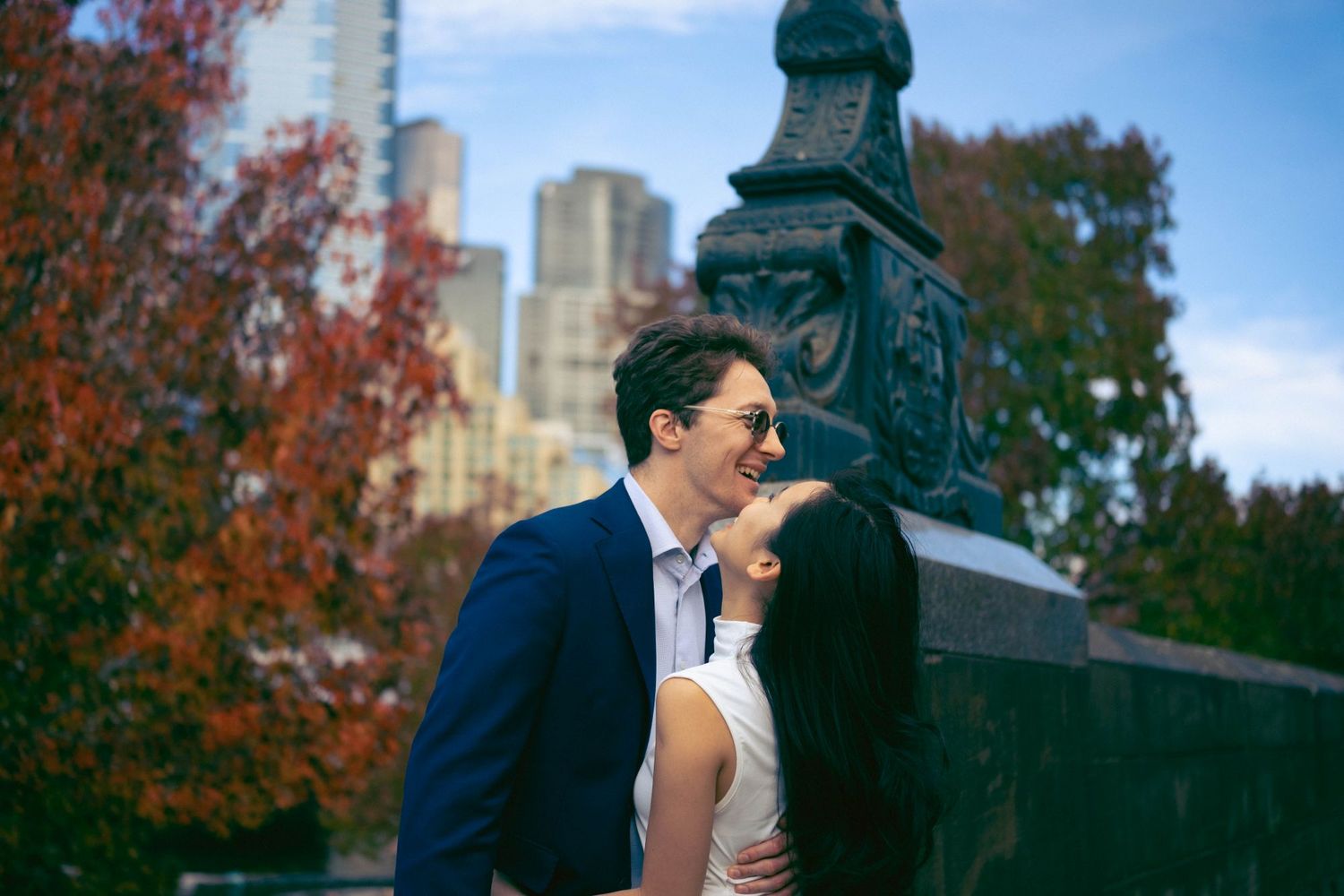 Romantic couple sharing an intimate moment near a decorative statue with autumn trees and city skyline behind them.