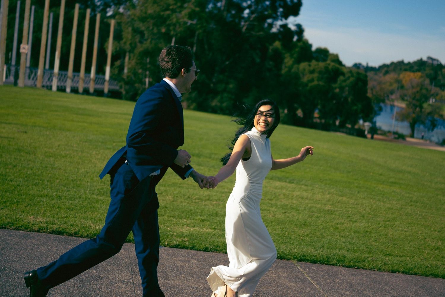 Couple in wedding attire holding hands and running across a green lawn with columns and a lake in the background.