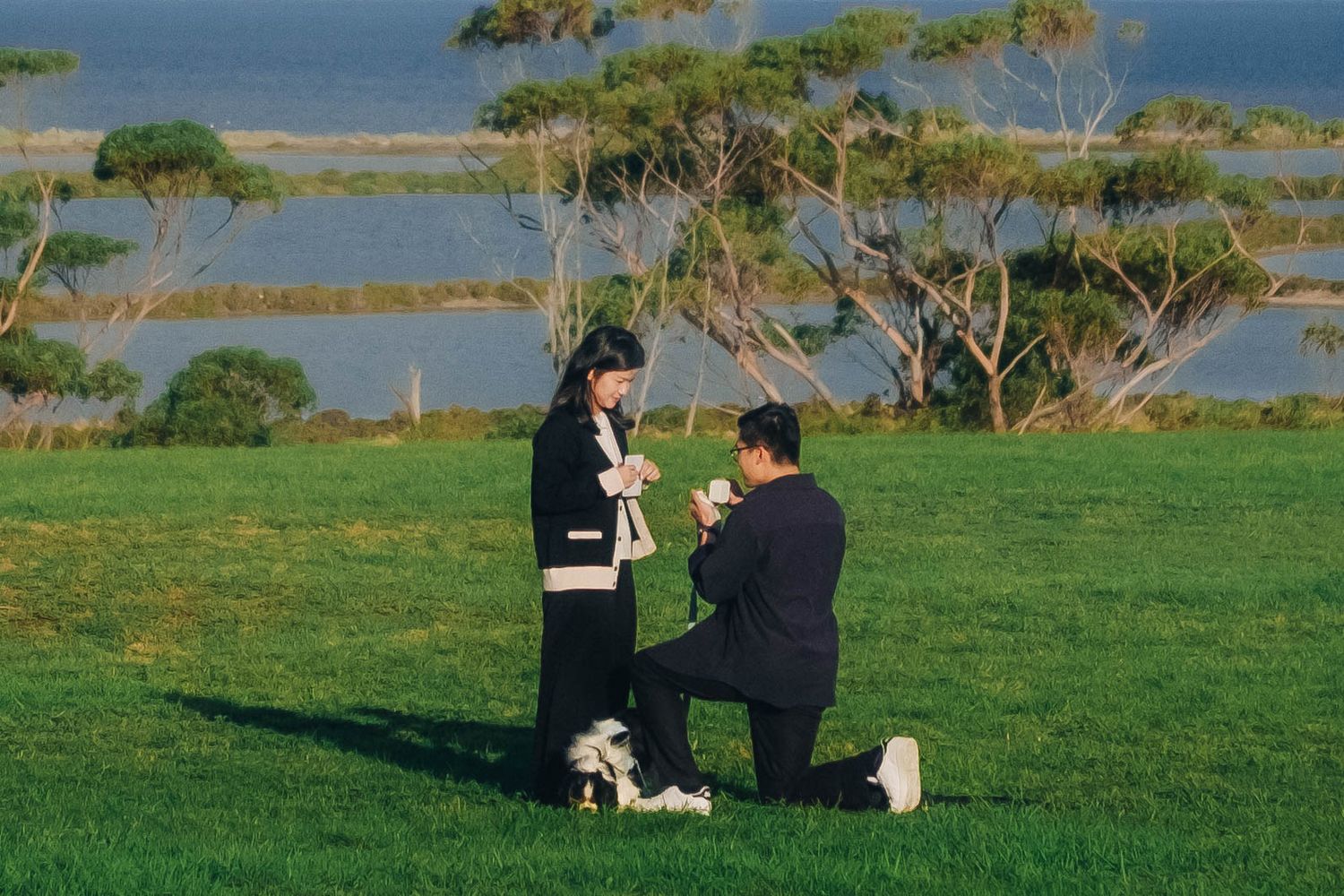 A man proposes on one knee to a woman on a green field with a scenic water view.