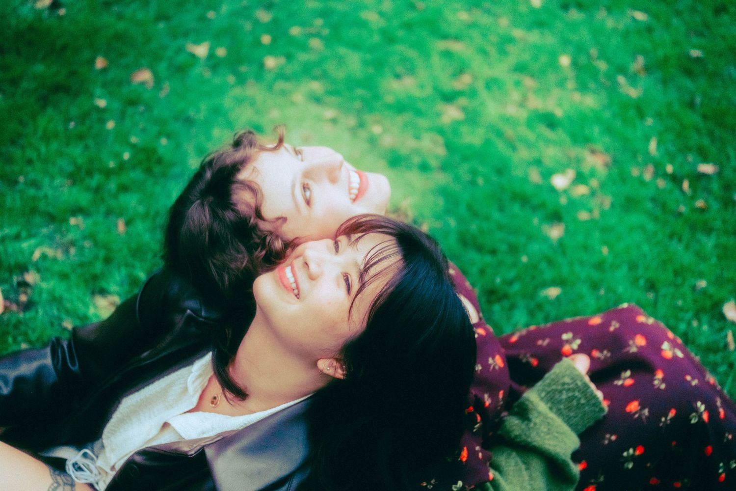 Two young women smiling and lying on green grass, enjoying a sunny day outdoors together.
