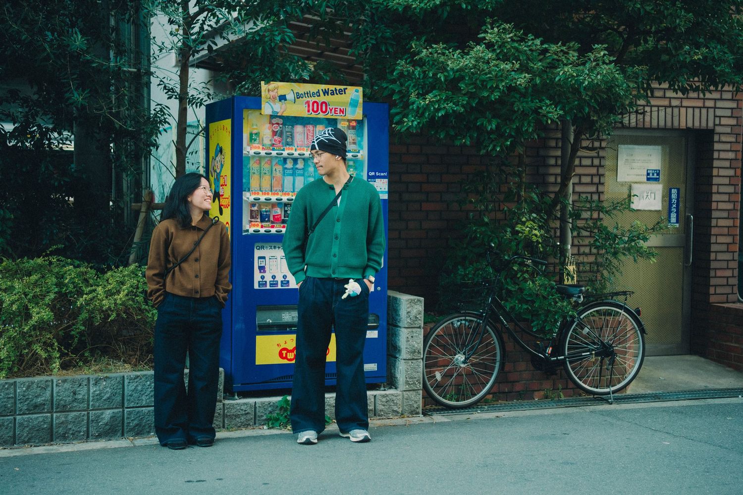 A couple stands talking next to a colorful Japanese vending machine on a tree-lined street.