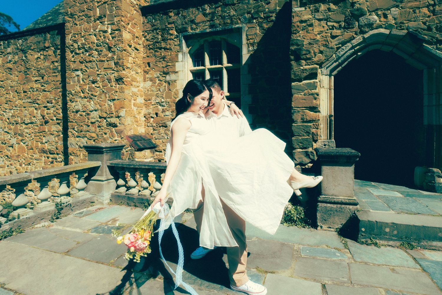 Groom carrying bride in white dress outside a rustic stone castle venue on their wedding day.