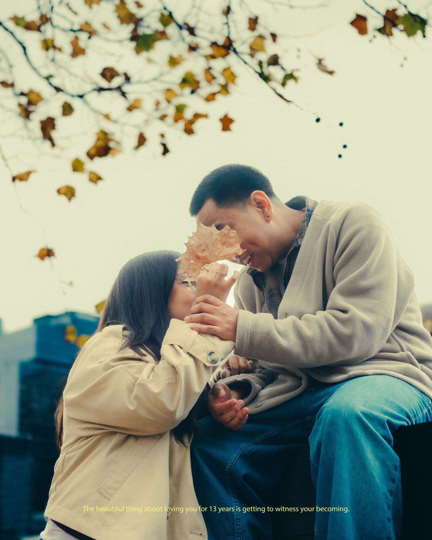 Romantic couple sharing a tender moment under autumn leaves, the man seated as the woman leans in close.