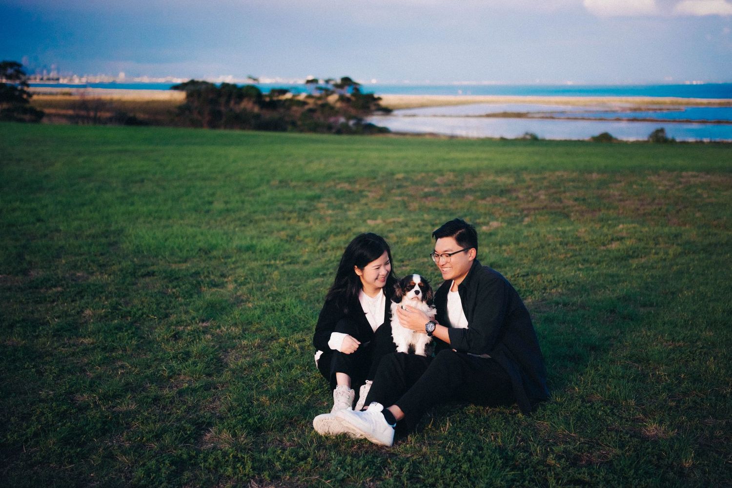 Couple dressed in black sitting on grass near a waterfront with a small dog between them.