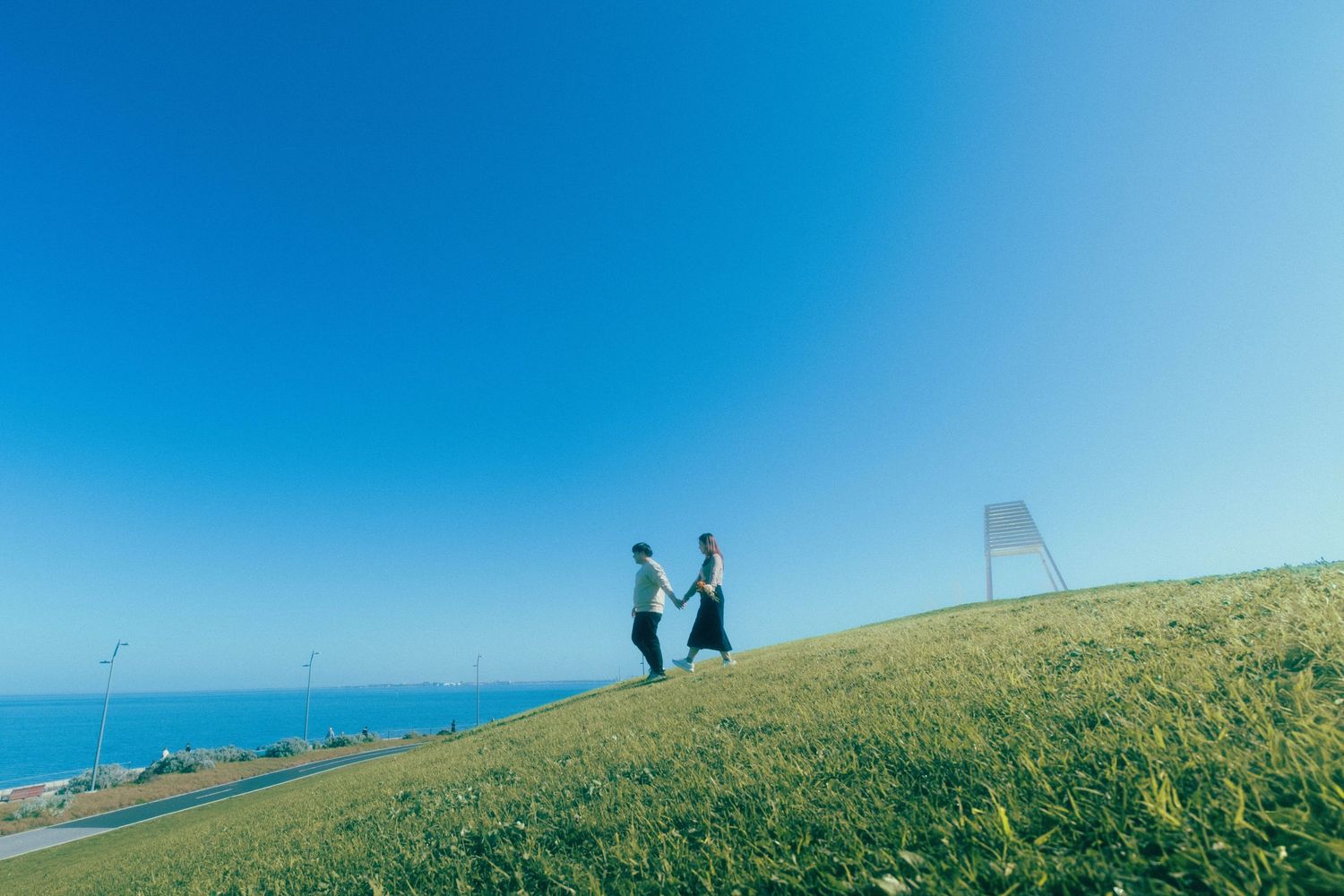 Couple holding hands walking on a green hillside with a clear blue sky and ocean view in the background.