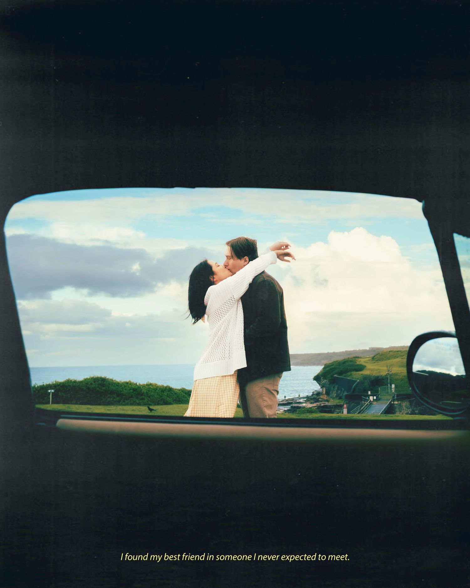Couple embracing and kissing viewed through car window with scenic ocean coastline and cloudy sky backdrop.