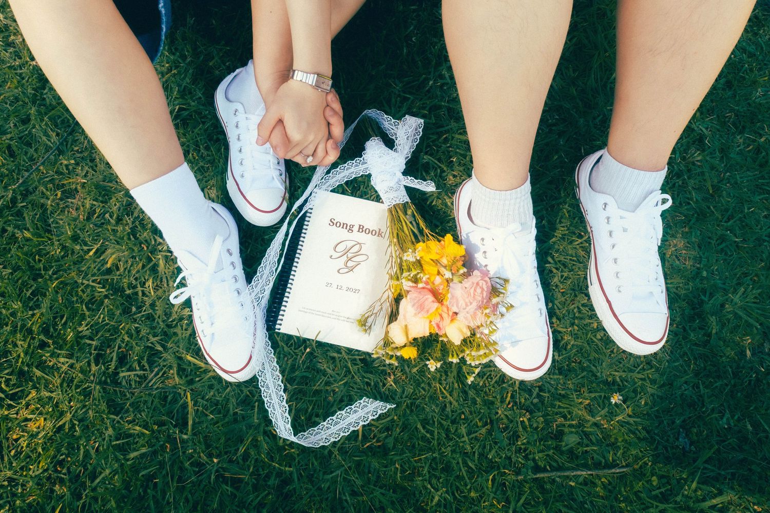 Two brides in white Converse sneakers hold hands over a floral bouquet and wedding program on green grass.