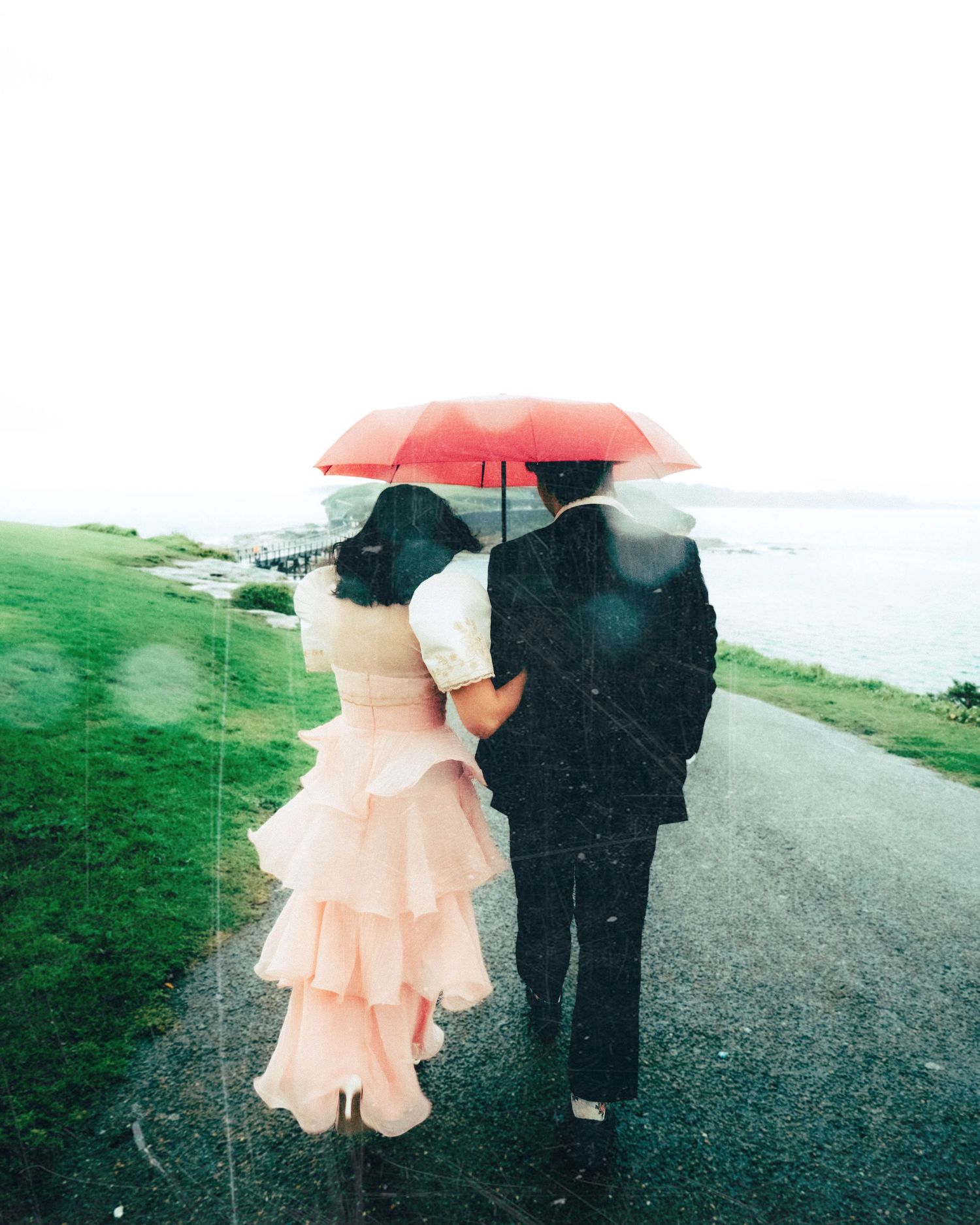 Couple sharing a red umbrella in the rain, walking along a coastal path, bride in pink ruffled gown.