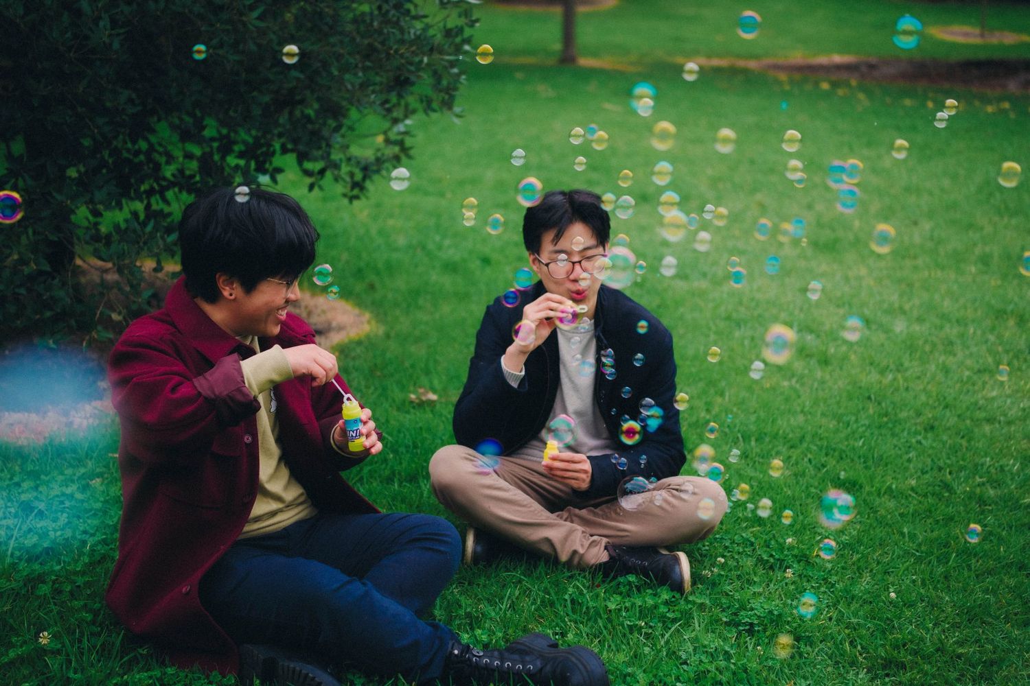 Two young men sitting on grass blowing colorful soap bubbles in a park.