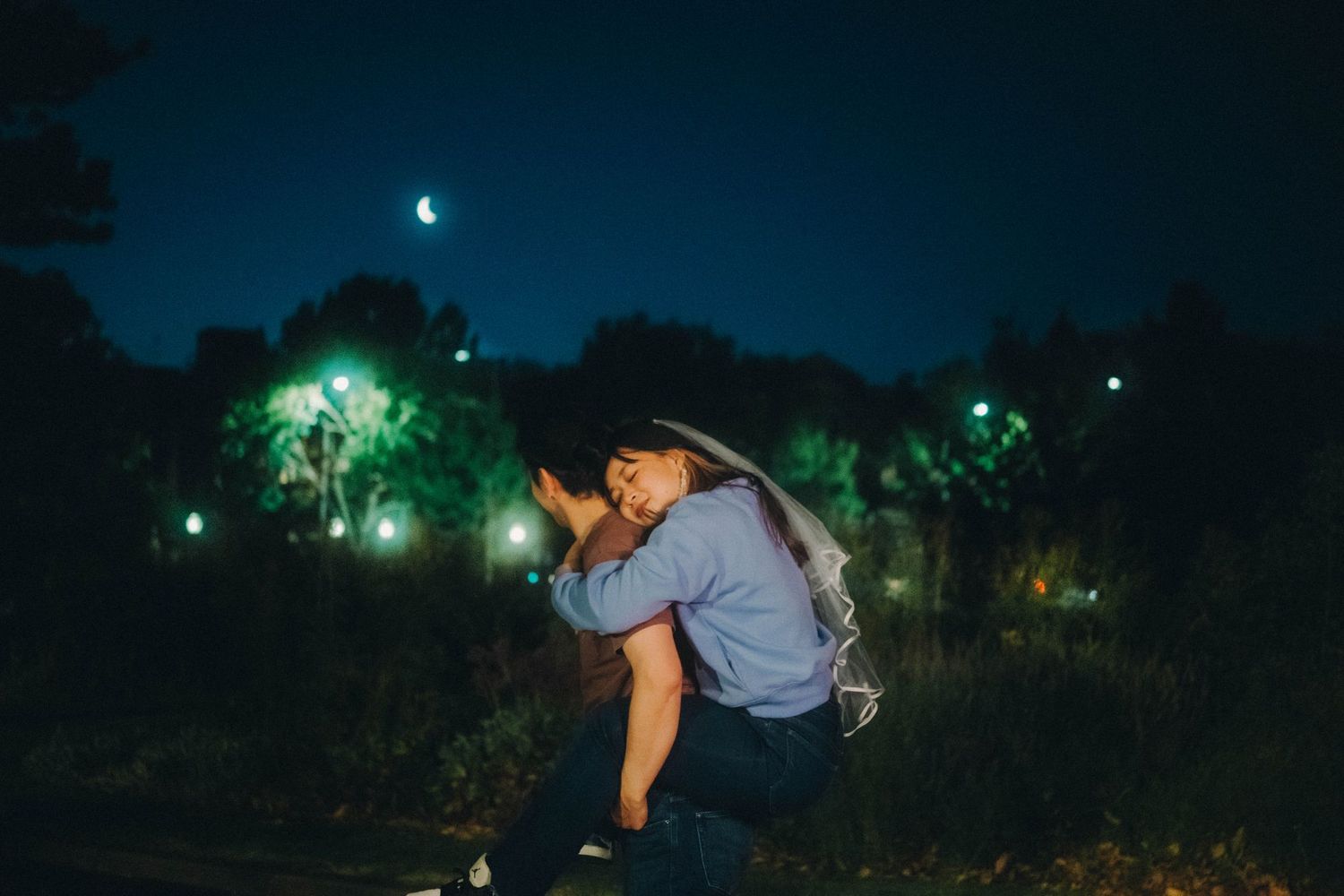 Young woman sitting outdoors at night under a crescent moon with glowing green lights in trees.