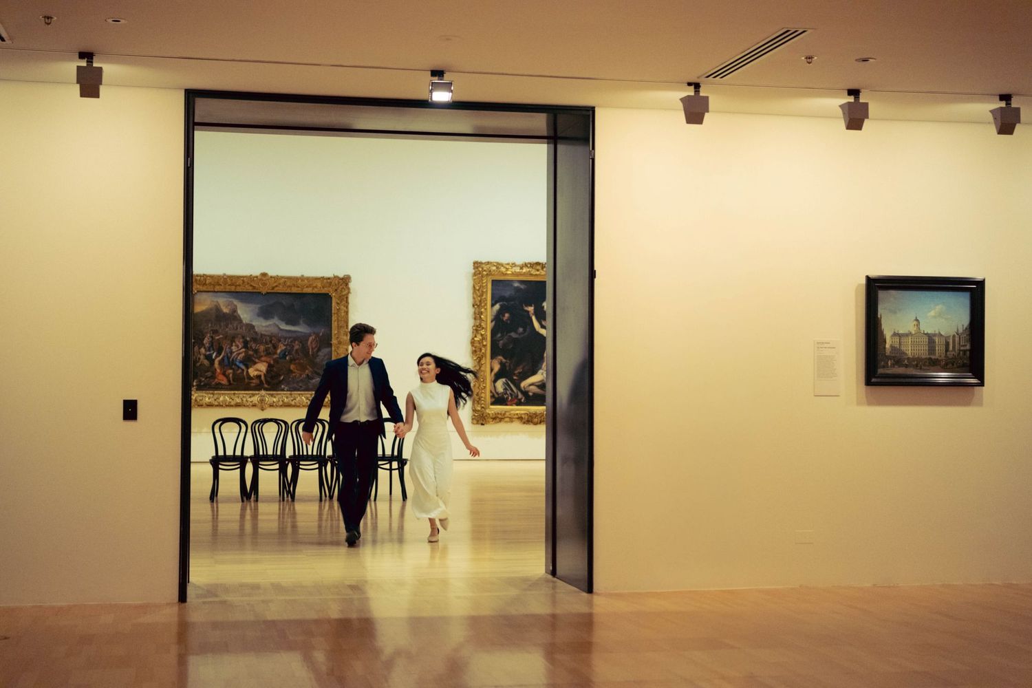 Couple running through an art museum gallery with classic paintings on walls.