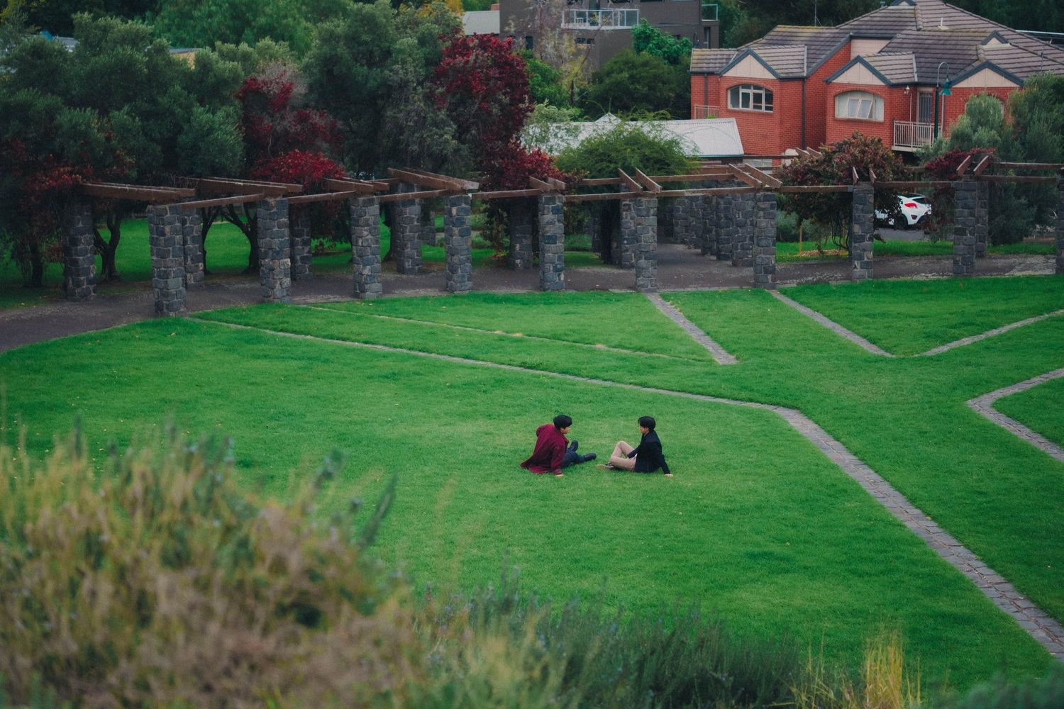 Two people sitting together on a green park lawn surrounded by colorful trees and stone pillars.