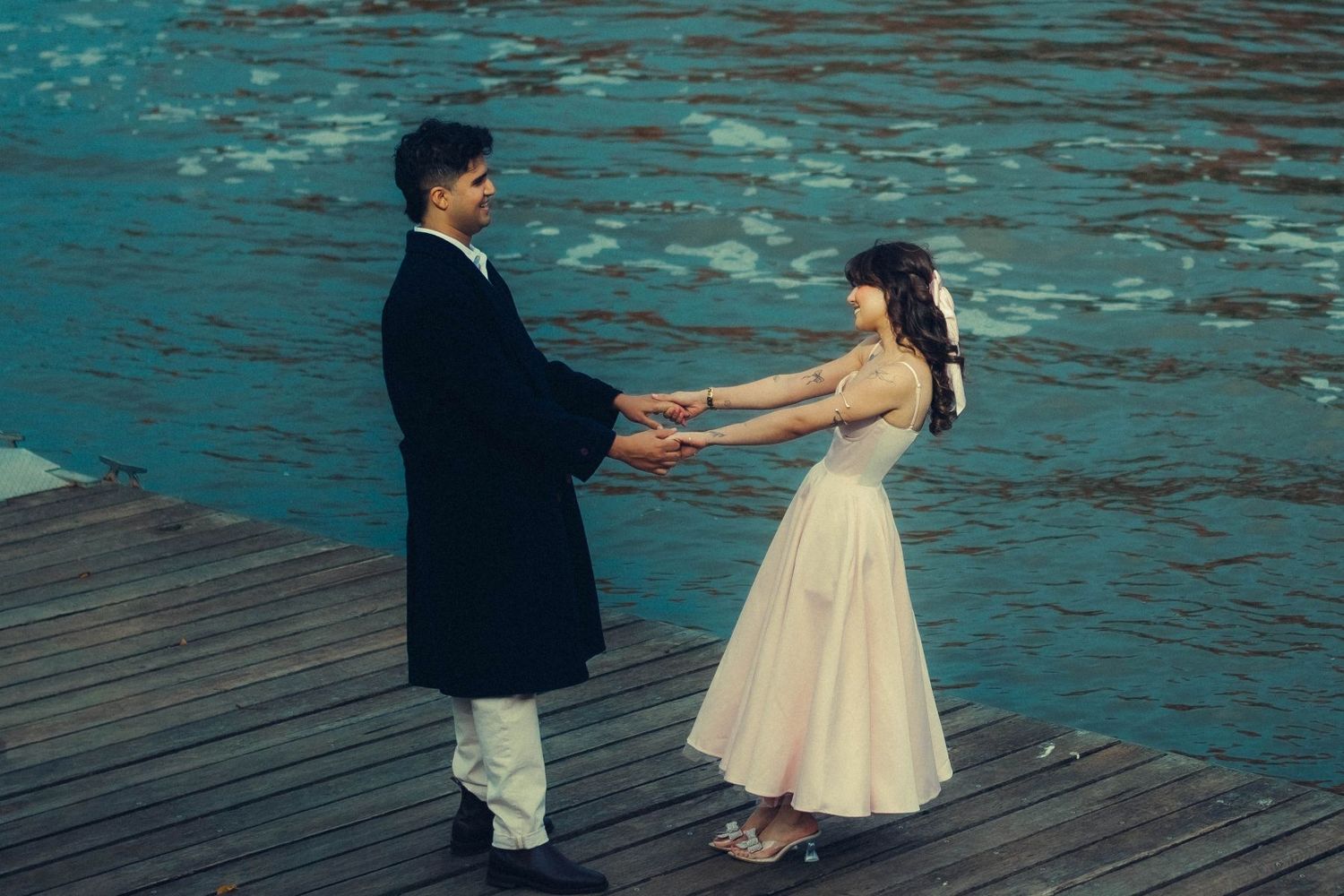 Couple holding hands on a wooden dock with blue water background.