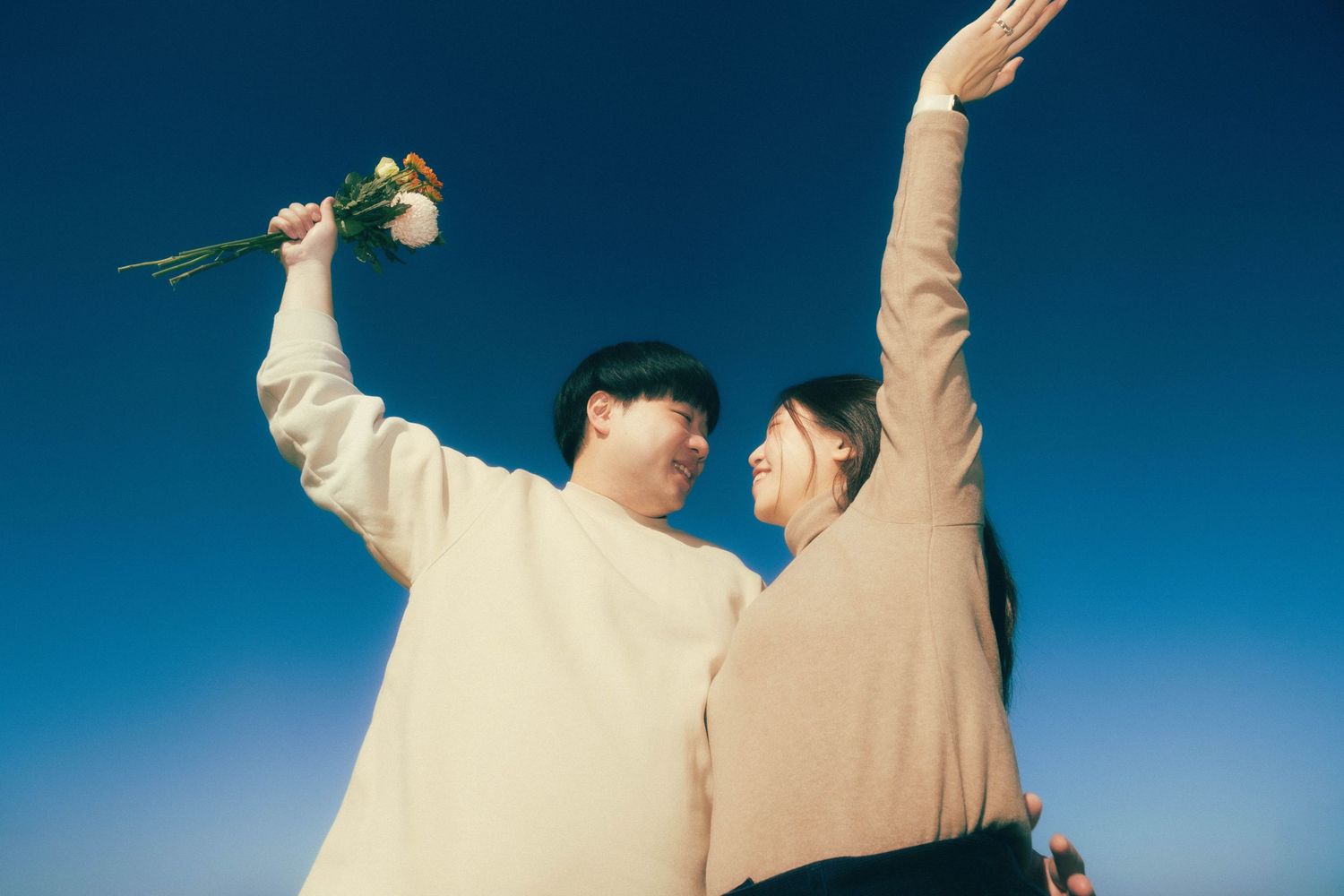 Couple holding flowers raising arms against blue sky in matching cream sweaters.
