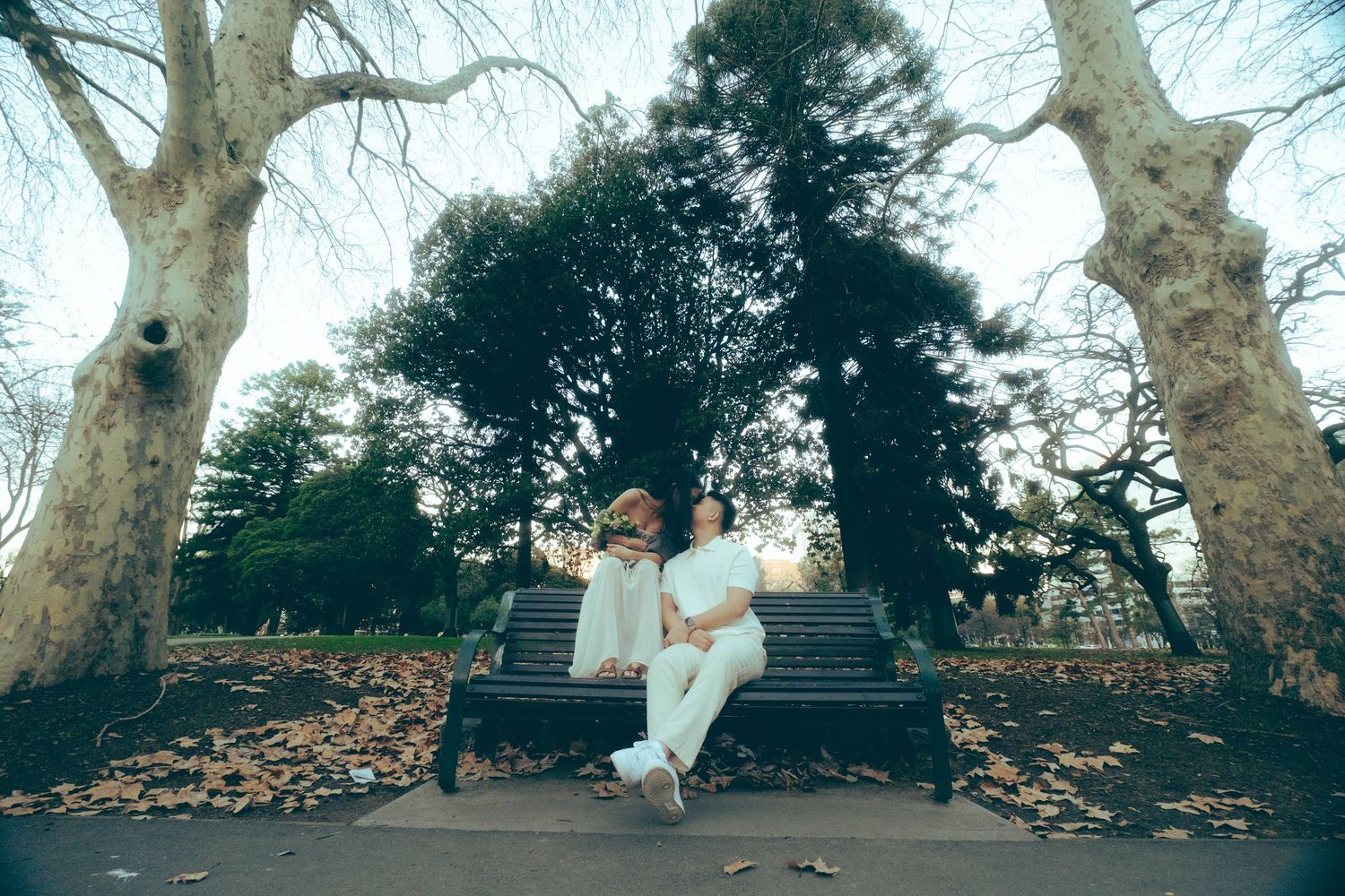Couple in white outfits sitting on a park bench surrounded by tall trees and autumn leaves.
