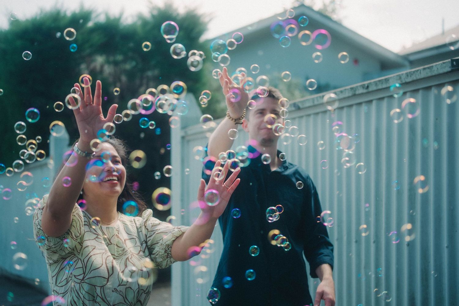 Three people laughing and reaching for colorful soap bubbles outdoors near a building.