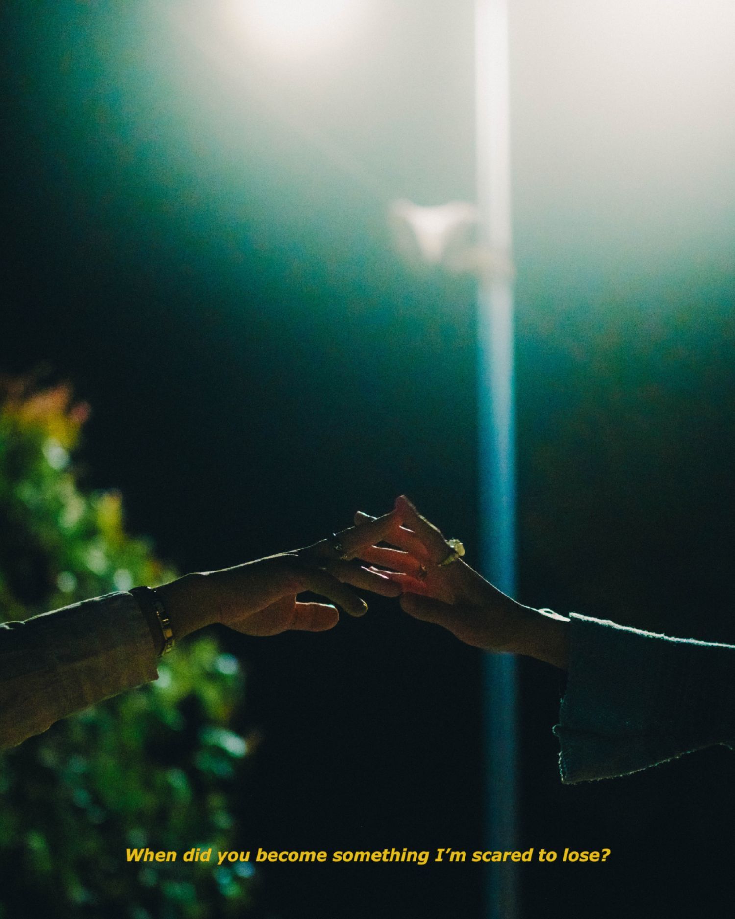 Two people reaching toward each other holding hands under a bright street light at night.