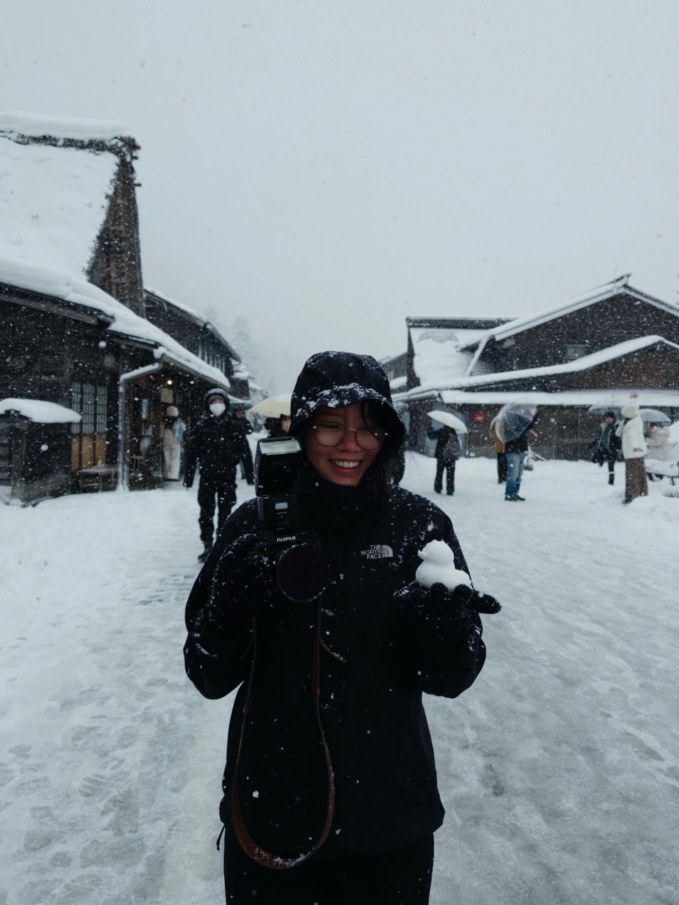A smiling woman in a black North Face jacket holds a snowball in a snowy Japanese village street.
