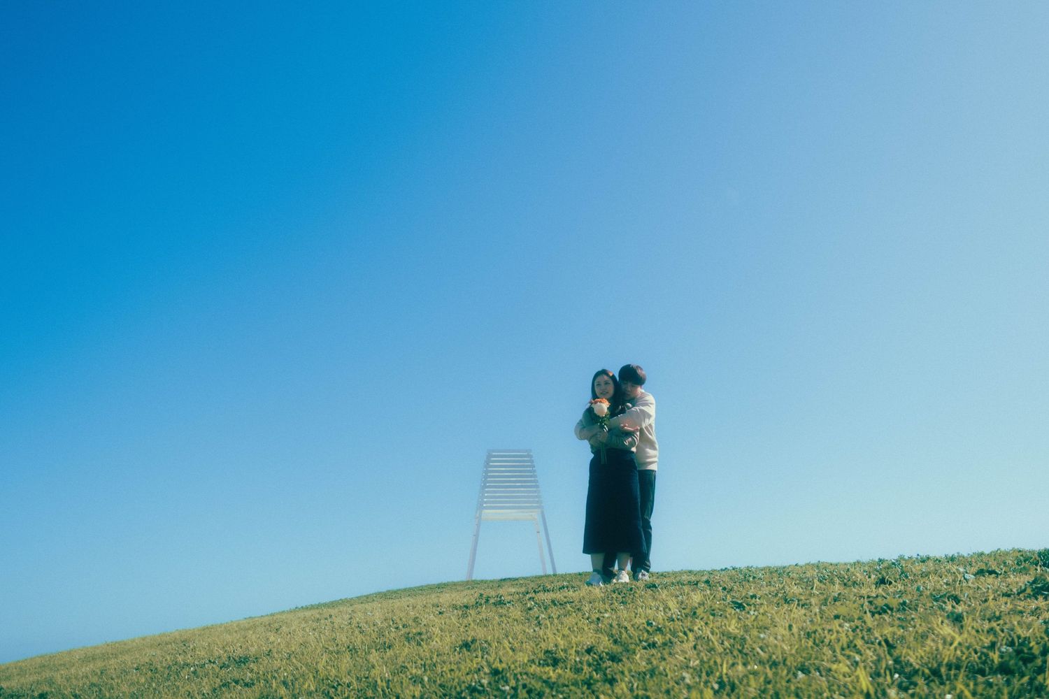Two people embracing on a green hillside under a clear blue sky, with a white chair standing nearby.