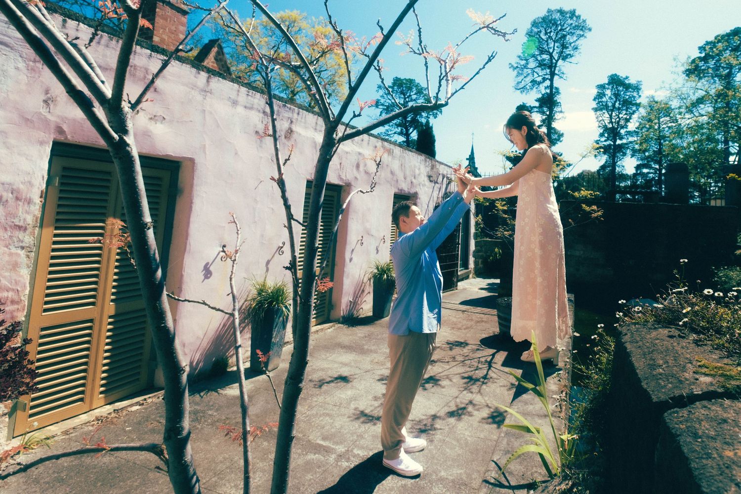Couple dancing outdoors near a pink building, woman in pink dress elevated, man in blue outfit holding her hands.