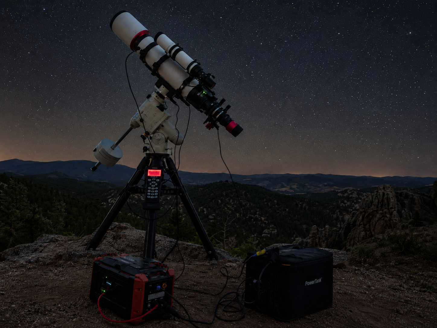 Astrophotography telescope on tripod atop rocky cliff with portable power station under starry night sky.