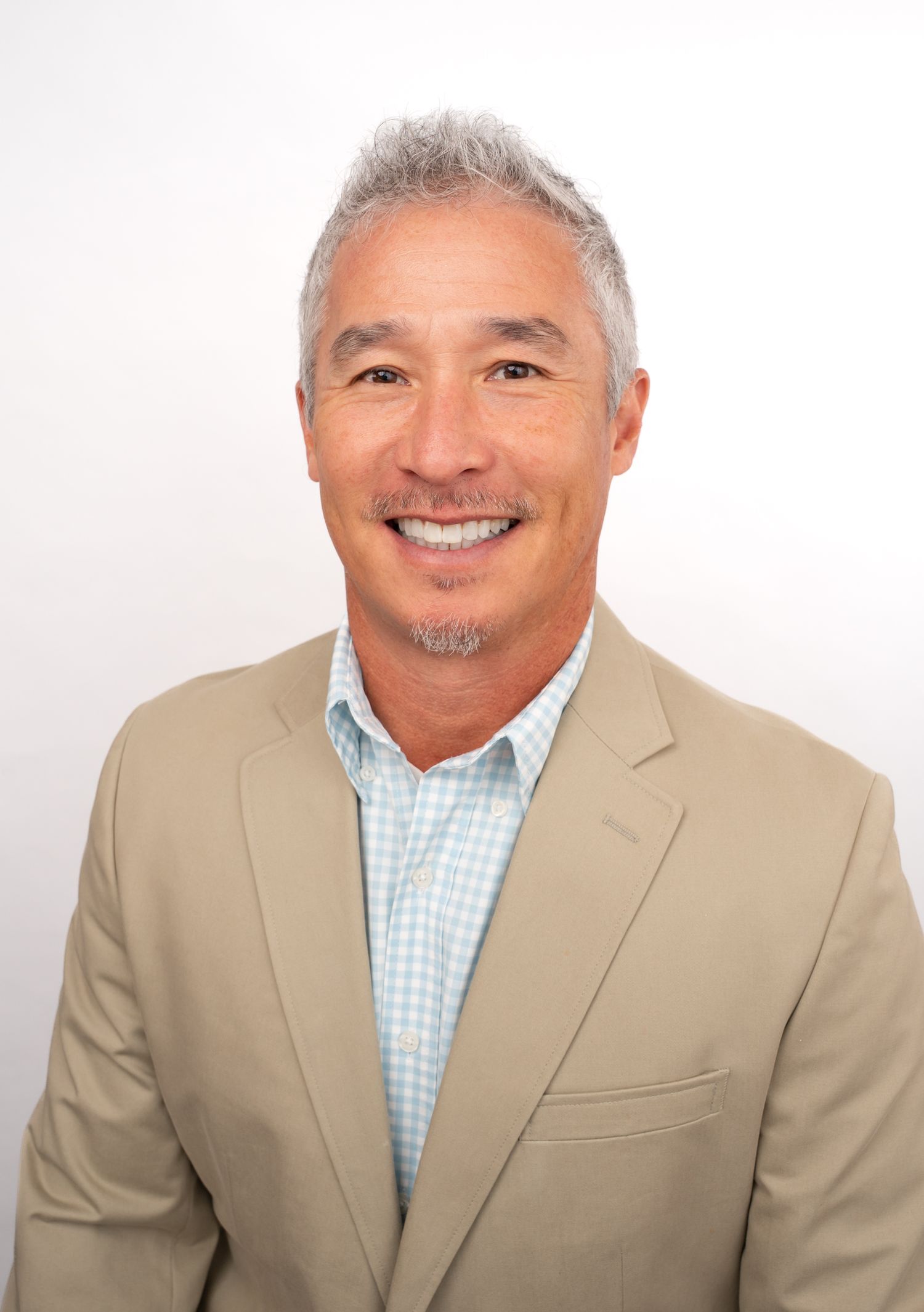 Smiling middle-aged man with gray hair wearing a tan blazer and light blue checkered shirt against a white background.