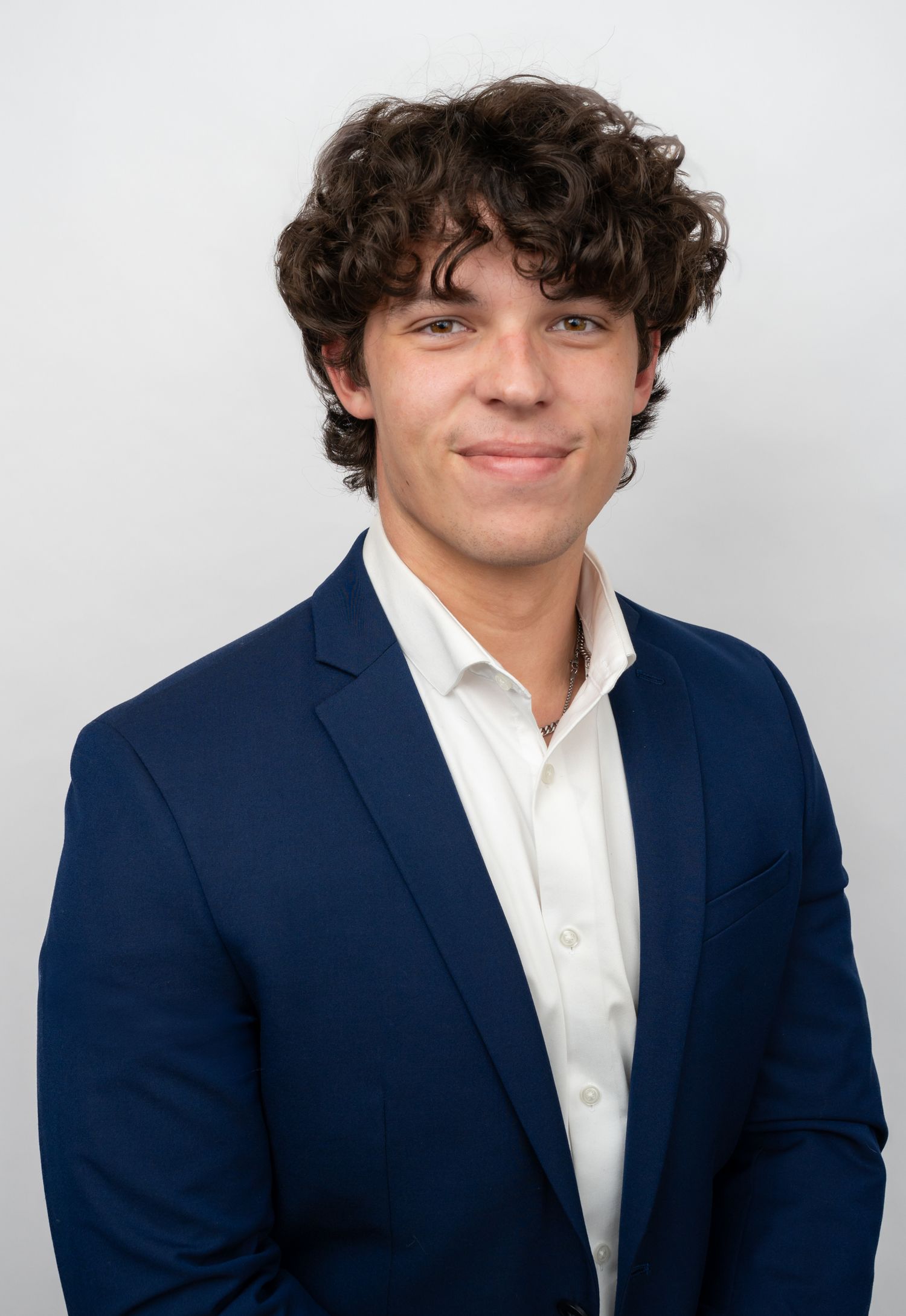 Young professional man with curly brown hair wearing a navy blue blazer and white shirt, smiling confidently.