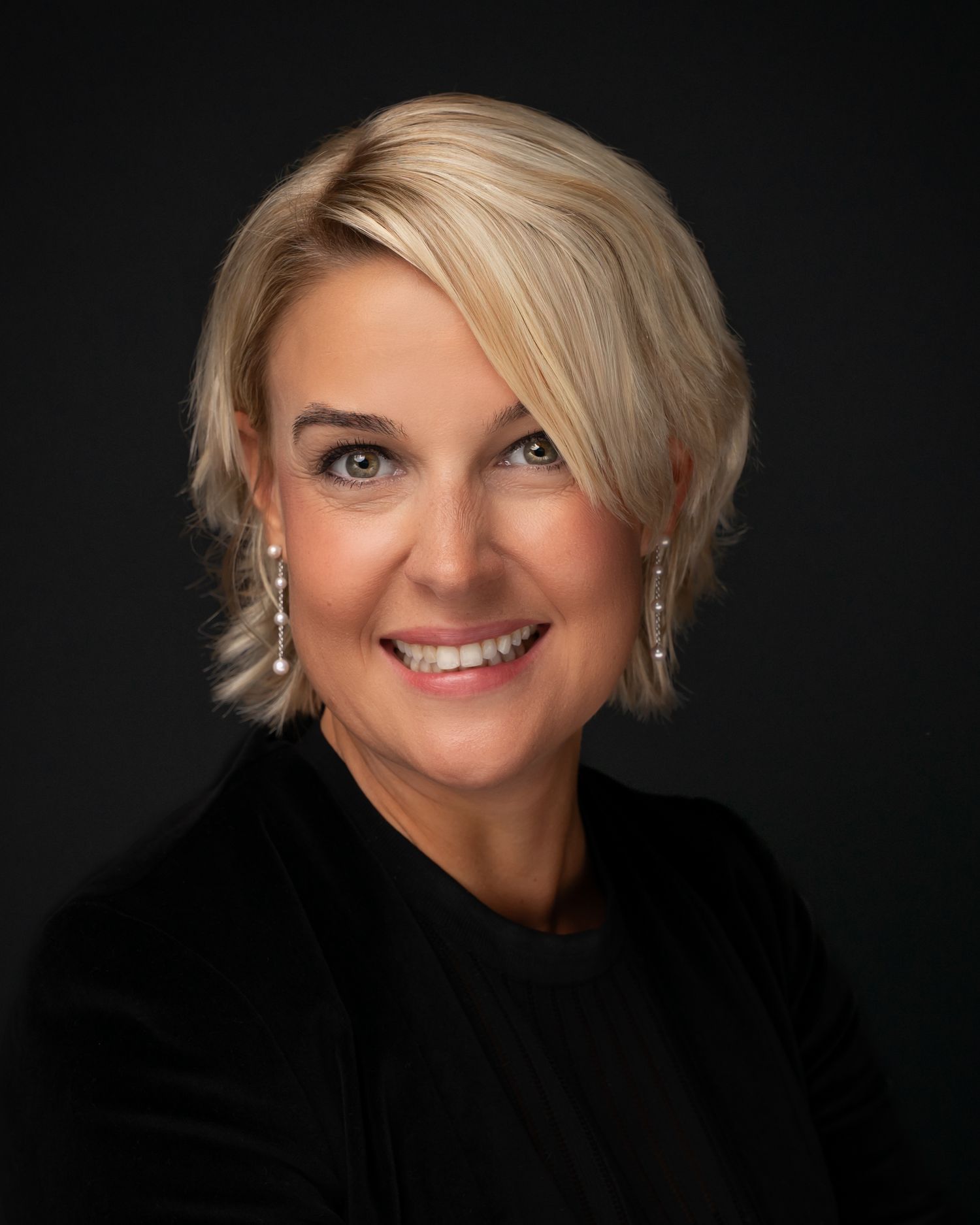 Professional headshot of a smiling blonde woman wearing black top and pearl drop earrings against dark background.