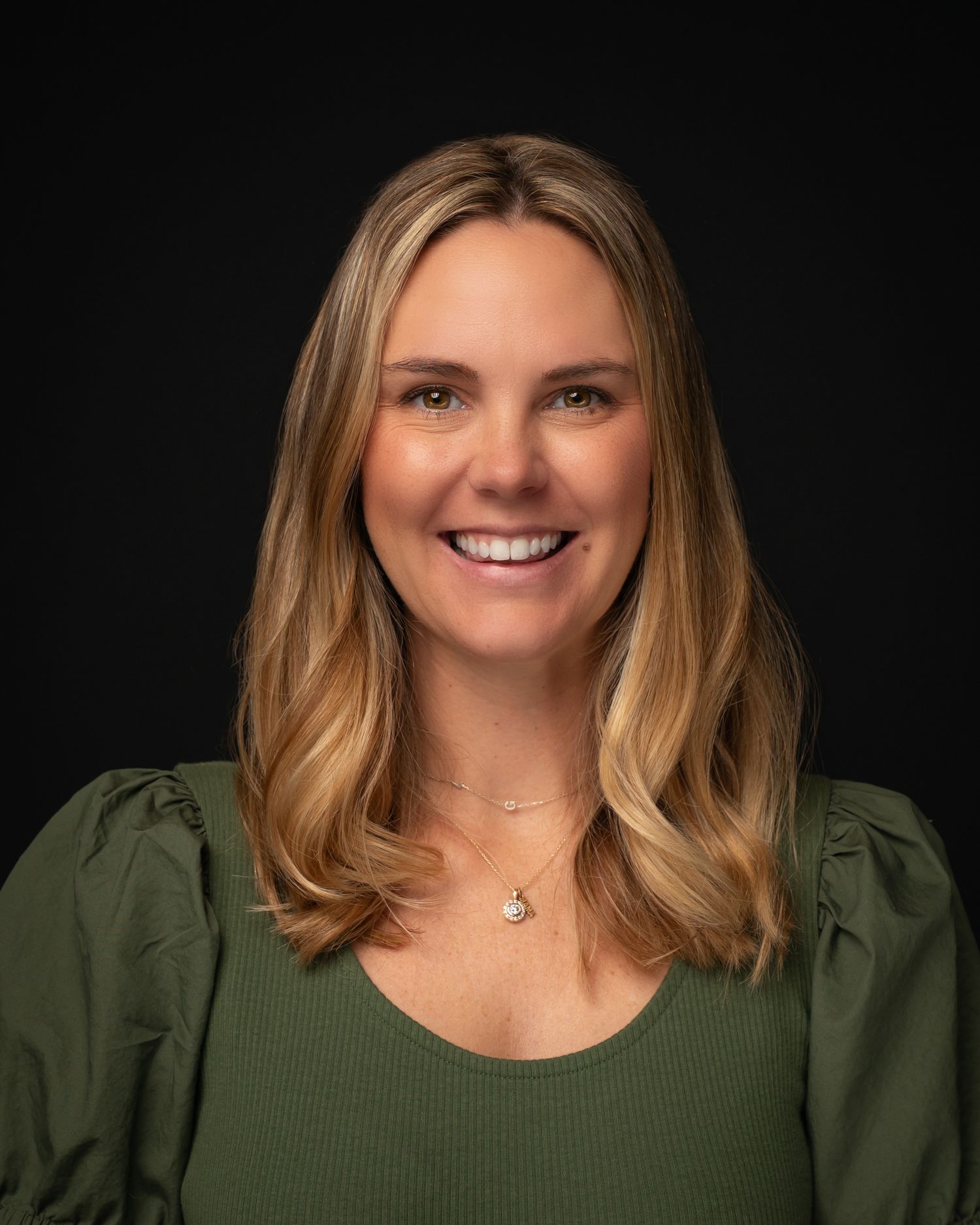 Smiling blonde woman wearing a green top and delicate necklace against a dark background.