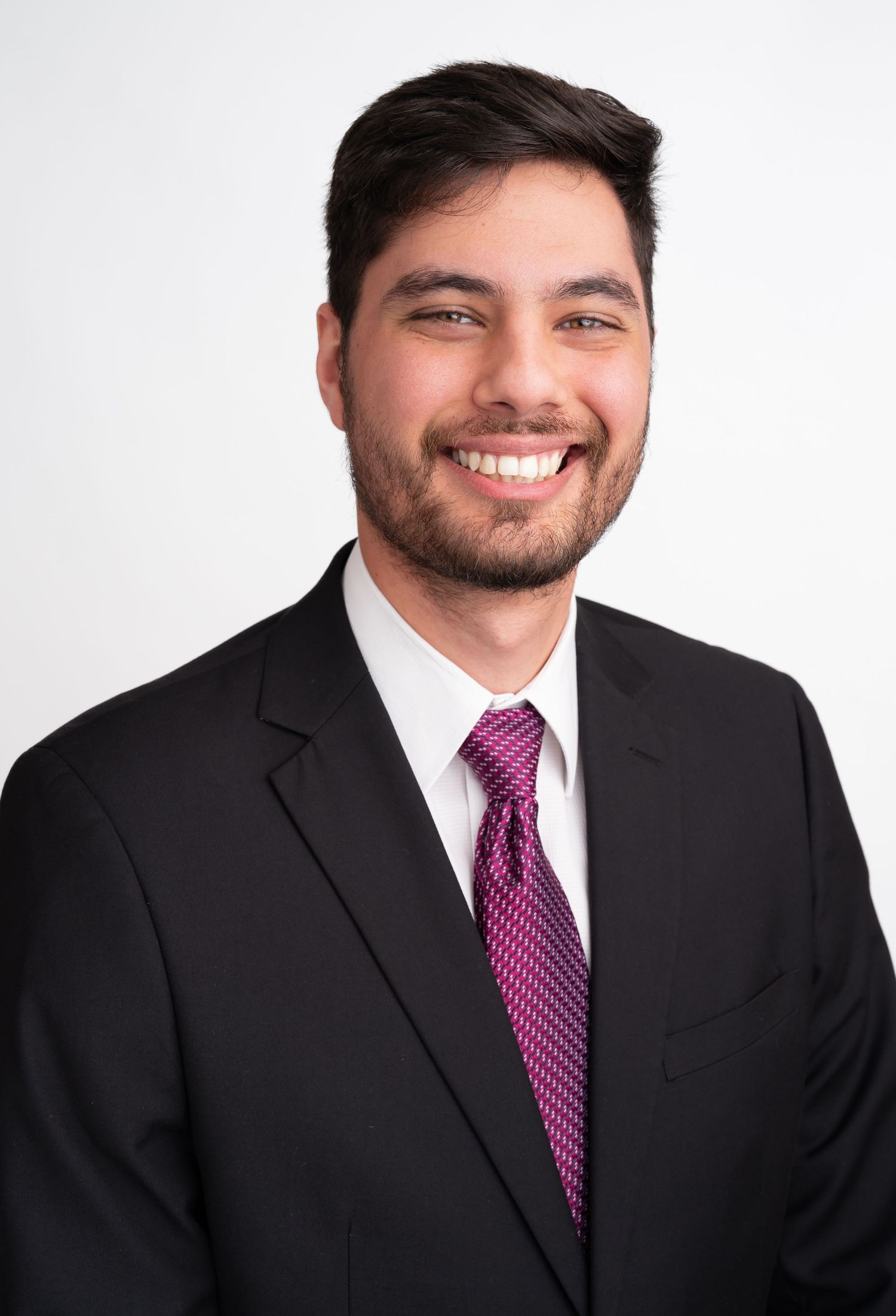 Professional headshot of a smiling young man with beard wearing a black suit and pink tie against white background.