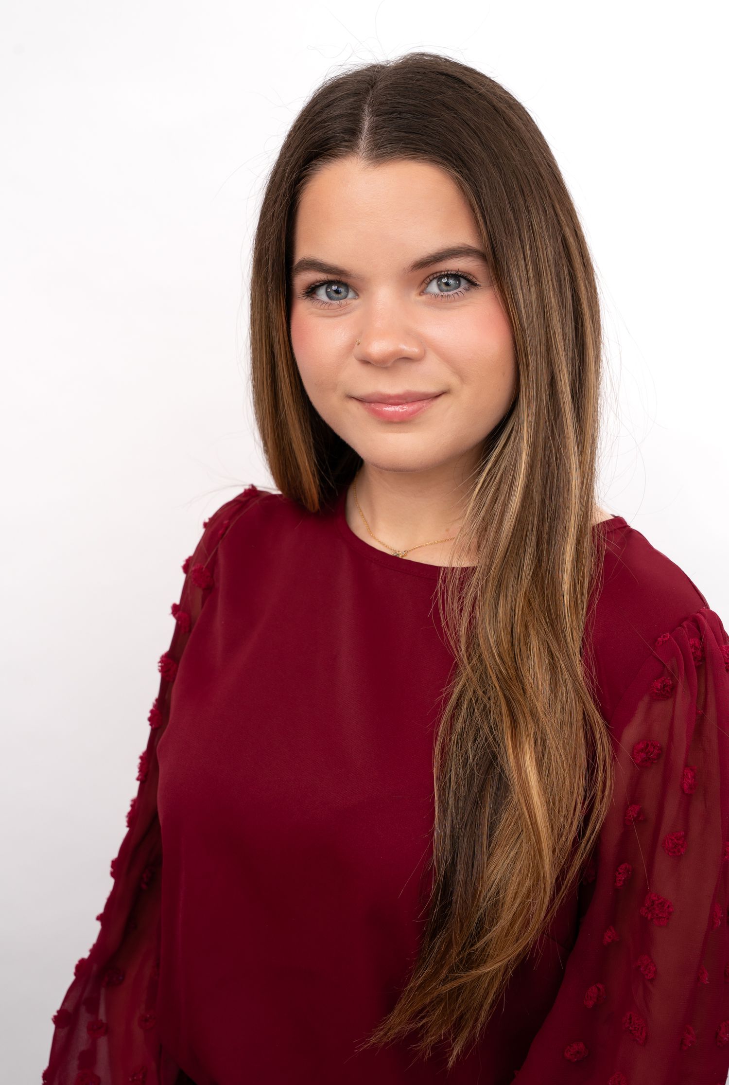 Young woman with long brown hair and blue eyes wearing a dark red ruffled blouse poses for a professional headshot.