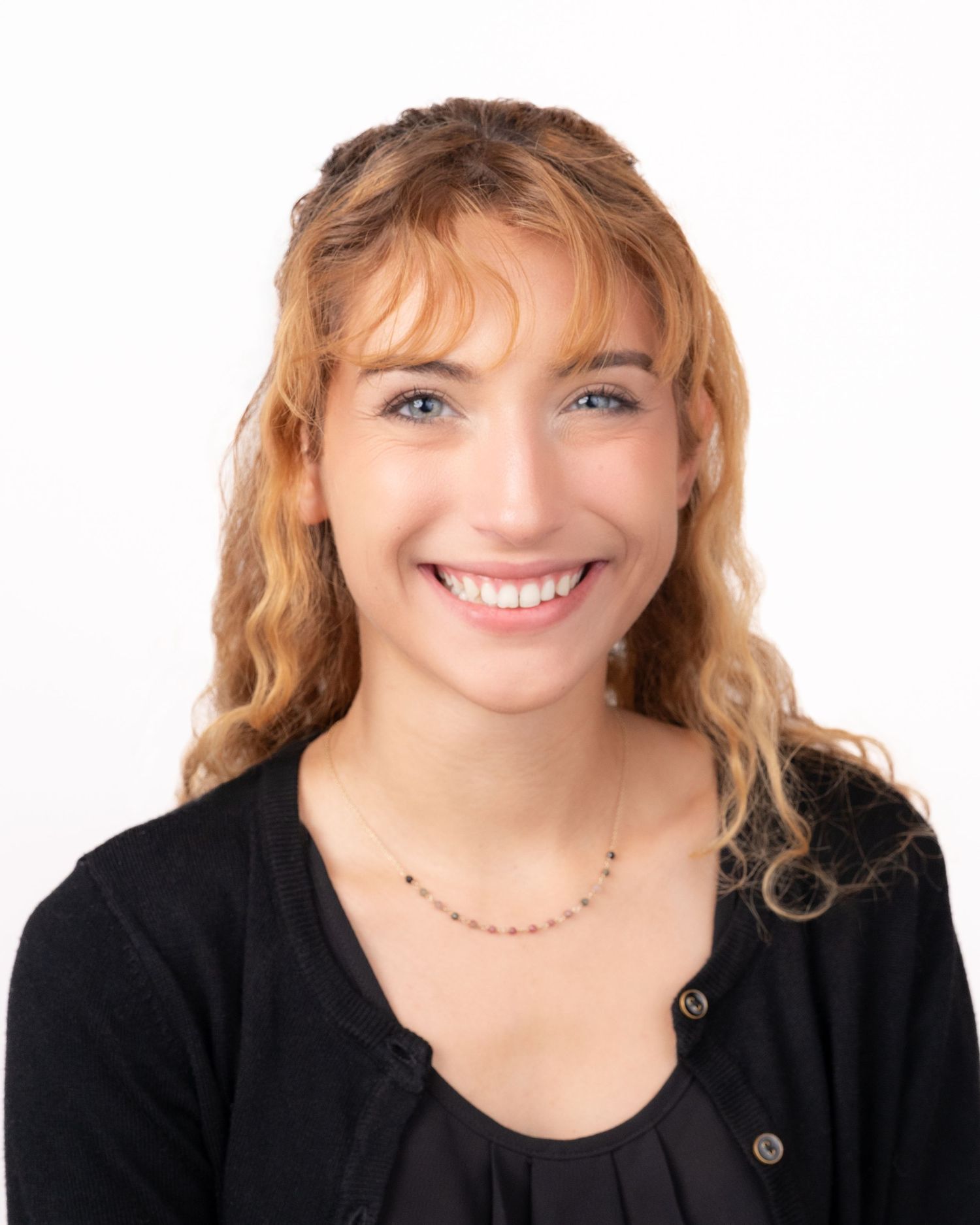 Smiling young woman with curly blonde hair and blue eyes wearing a black cardigan and delicate necklace.