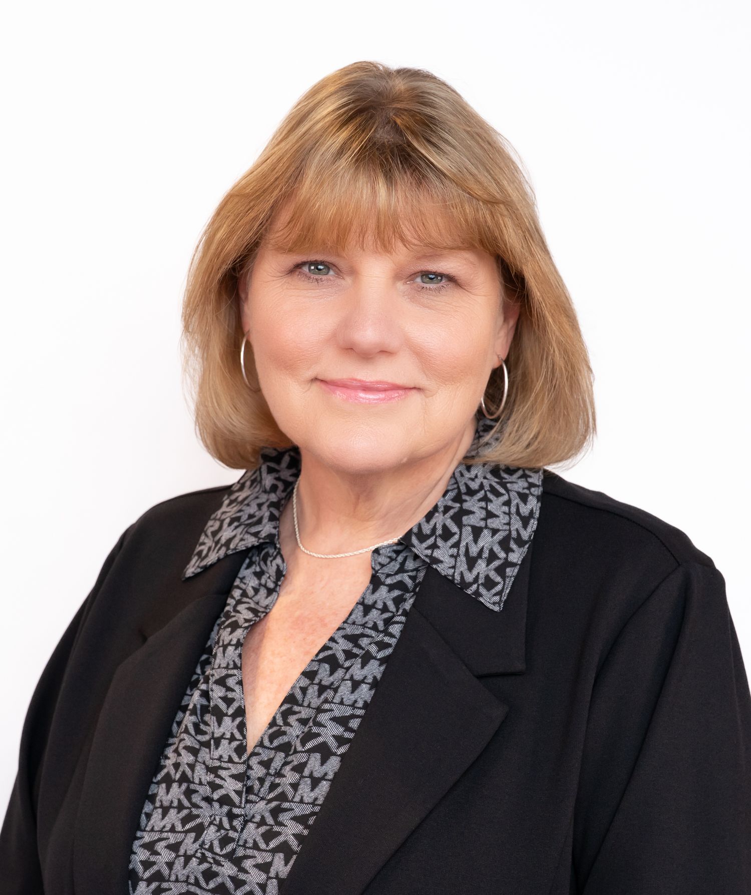 Professional headshot of a smiling middle-aged woman with blonde hair wearing a black blazer and patterned blouse.