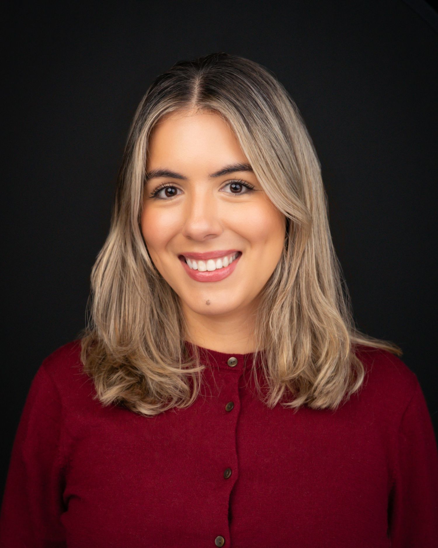 Young woman with blonde hair smiling, wearing a red cardigan, professional headshot on dark background.