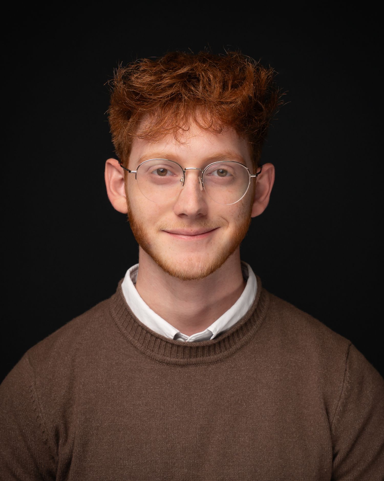 Young man with curly red hair and glasses wearing a brown sweater over a white collared shirt.